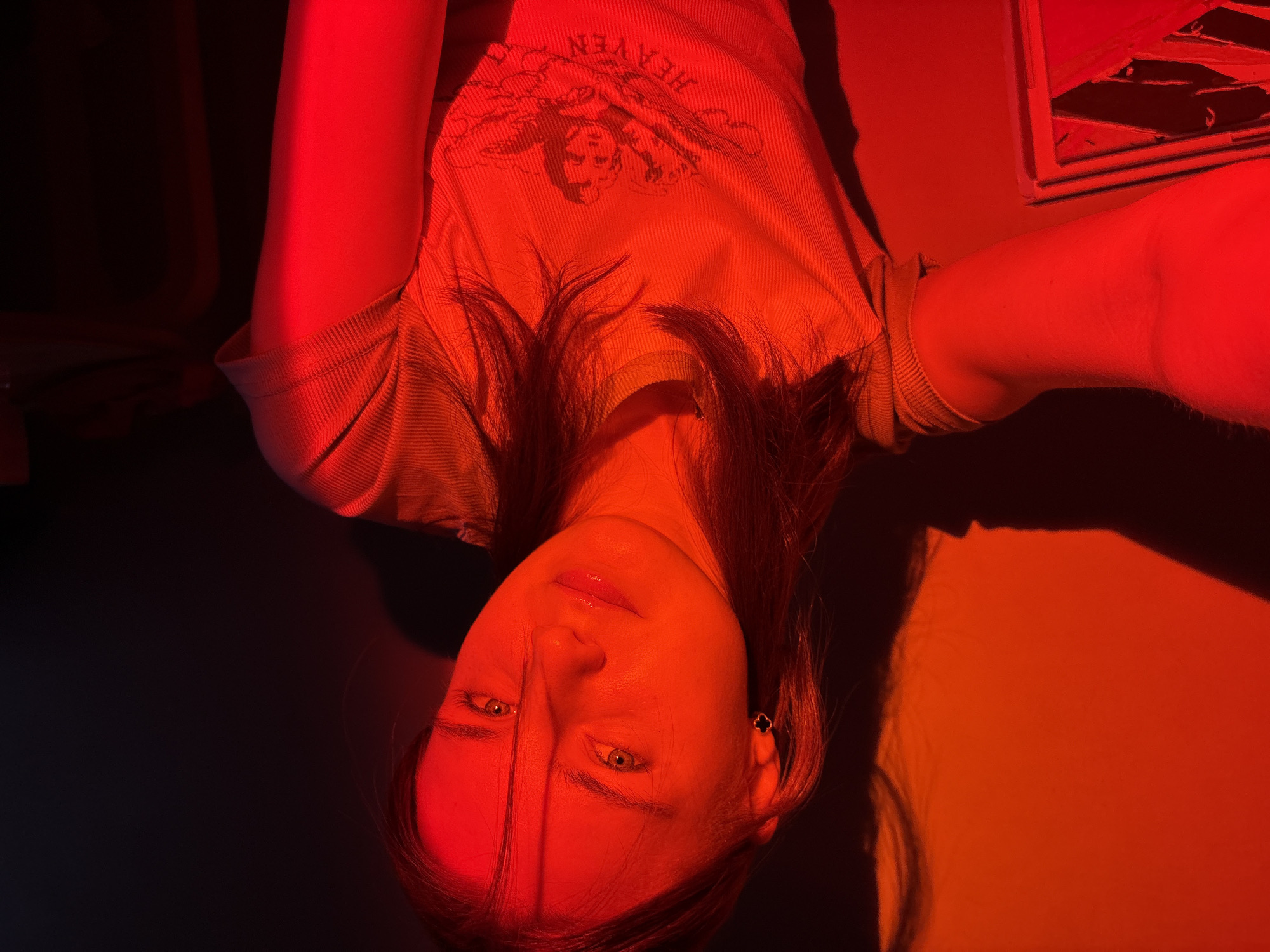 The image presents a close-up, stylized portrait of a person, likely a young woman, lying down and looking towards the camera. The entire scene is drenched in a vivid, intense red light, creating a dramatic and high-contrast aesthetic.
The subject has long, dark hair, with some strands falling across her face. Her eyes appear light-colored, possibly green or hazel, and her expression is neutral to slightly contemplative. She is wearing a light-colored, ribbed t-shirt, which is strongly illuminated by the red light. A distinct graphic on the t-shirt shows cherubic figures and the word "HEAVEN" printed upside down relative to the subject's orientation. A dark, possibly clover-shaped earring is visible on her right ear.
The activity appears to be posing for a photograph, likely a selfie, within a controlled lighting environment indoors. The pervasive red light casts deep shadows on parts of her face and the background, while highlighting her features and the texture of her clothing. In the upper right corner of the image, there's a reflection or a glimpse of an object with abstract, angular shapes in red and black, possibly a screen or a piece of art.
The location is unknown, Russia. The artificial lighting makes it impossible to determine the time of day or weather conditions. The overall mood is artistic, intense, and somewhat mysterious due to the striking color palette and lighting.