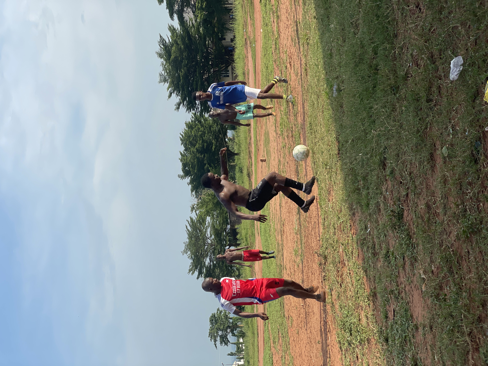 The image depicts an outdoor soccer game or practice session involving at least five men on a sunny day in Ogbogbo, Nigeria. The setting is a rustic, uneven field composed of reddish-brown dirt patches and green grass.
In the foreground, a man wearing a red and white jersey with "Emirate FLY BETTER" visible text stands to the left, facing right. Behind him, another man in red shorts is visible. The central action features a shirtless man in black shorts and socks, captured mid-air performing an acrobatic overhead kick (bicycle kick) with his right leg, aiming for a white soccer ball floating in front of him. Further back, another shirtless man in light green shorts, possibly with the numbers "68" or "89" on them, is standing. To the right, a man in a blue and white jersey, with "NEW POLY" partially visible, and white shorts is standing, observing the play.
The background is lined with lush green trees, and a faint structure of a building can be seen through them. The sky is bright and clear with scattered white clouds, indicating broad daylight and fair weather. The players appear focused on the game.
