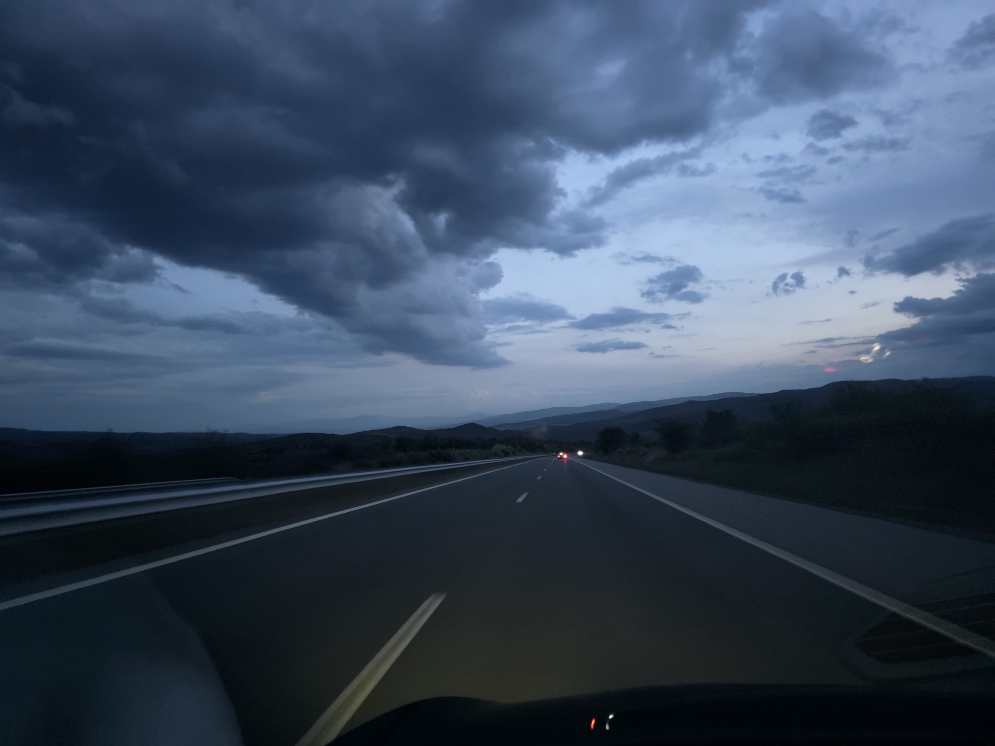 The image presents a first-person view from inside a vehicle traveling on a multi-lane highway at dusk or twilight. The dominant feature is the dramatic, heavily overcast sky, filled with dark, dense clouds in shades of deep gray and blue. Patches of lighter blue are visible where the cloud cover is less thick, and a faint reddish-pink glow can be seen near the horizon on the right, suggesting the last remnants of a sunset.
The asphalt highway stretches into the distance, clearly marked by white dashed and solid lines, and appears somewhat reflective due to the low light. A metal guardrail runs along the left side of the road. In the mid-ground, red taillights from several distant vehicles are visible on the highway, indicating other traffic. The landscape surrounding the road consists of dark, undulating hills and mountains, which appear silhouetted against the dimming sky, suggesting a rural or mountainous setting.
The foreground shows the top portion of the car's dashboard and windshield, with a soft glow from the vehicle's headlights illuminating the immediate road ahead. No people are visible. There is no discernible text within the image. The scene captures a journey through a quiet landscape under a moody, darkening sky, consistent with the Blatino, Bulgaria location context.