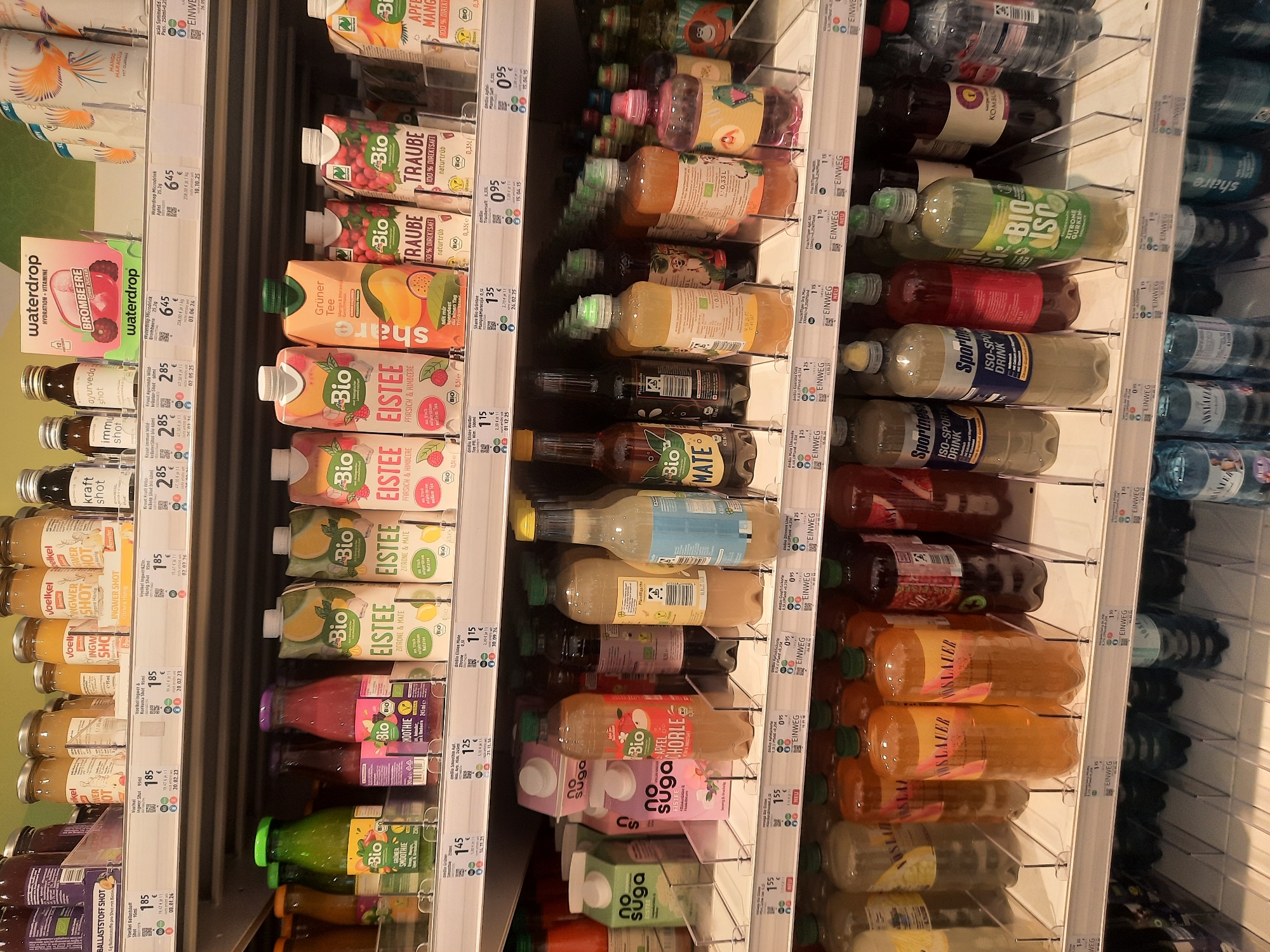 The image shows a retail display of beverages in a grocery store. The shelves are stocked with various drinks including juices, teas, smoothies, and water bottles. The products are primarily packaged in cartons and plastic bottles. Some of the brands visible include "dmBio," "Share," "Vöslauer," and "Sportness." The pricing information is displayed on shelf tags, with prices ranging from 0.95€ to 1.85€. The lighting is typical of a supermarket, with overhead fluorescent lights illuminating the products. The shelves are made of metal with plastic dividers. In the background, other shelves with more products are visible. The scene appears to be a typical supermarket aisle. This image was taken at a dm-drogerie markt in Rosenheim, Germany.