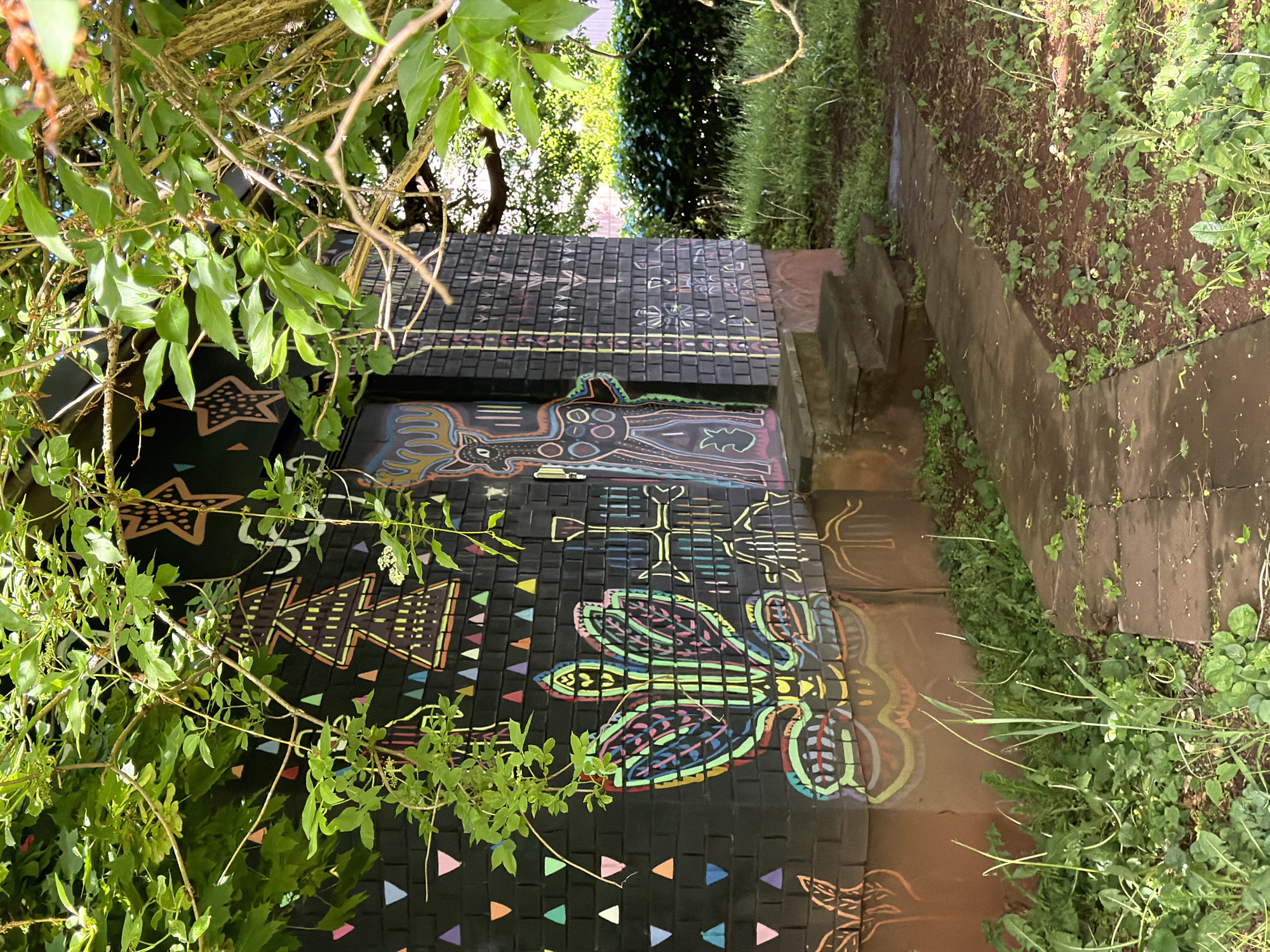 The image shows a set of outdoor stairs in Esch-sur-Alzette, Luxembourg, decorated with colorful, stylized artwork on black tiles. The artwork features diverse motifs, including a deer-like creature, stylized birds, and geometric patterns. The stairs lead downwards, flanked by greenery and a small pathway covered in vegetation. The scene is partially obscured by tree branches and leaves in the foreground.