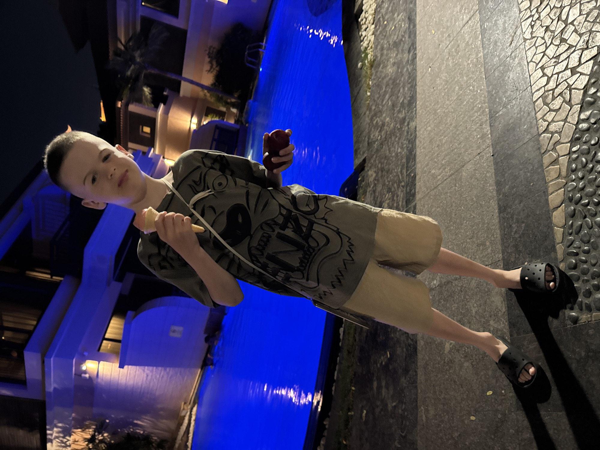 A young male child, estimated to be between 8 and 12 years old, stands on a paved walkway at night. He has a short, neatly trimmed haircut and looks directly at the camera with a neutral expression. He is wearing a dark olive-green short-sleeved t-shirt with white graphic designs, light tan shorts, and black slide sandals. In his right hand, he holds a light pink or peach-colored ice cream cone. In his left hand, he holds two small, round red fruits. A light-colored strap crosses his chest.
Behind the child, a vivid blue swimming pool is brightly illuminated from within, with reflections visible on its surface. The pool is bordered by small, light-colored decorative stones. In the background, resort-style buildings with warm-toned lighting are visible, featuring windows emitting light and architectural details. Palm trees are present among the buildings and dark foliage. The setting suggests a luxurious outdoor environment at night. The paved walkway beneath the child features distinct sections of dark grey, lighter grey, and a decorative mosaic of irregular light-colored stones and dark pebbles. The architectural style, prominent palm trees, and luxurious resort-like features align with common aesthetics found in high-end developments in Dubai, United Arab Emirates.
