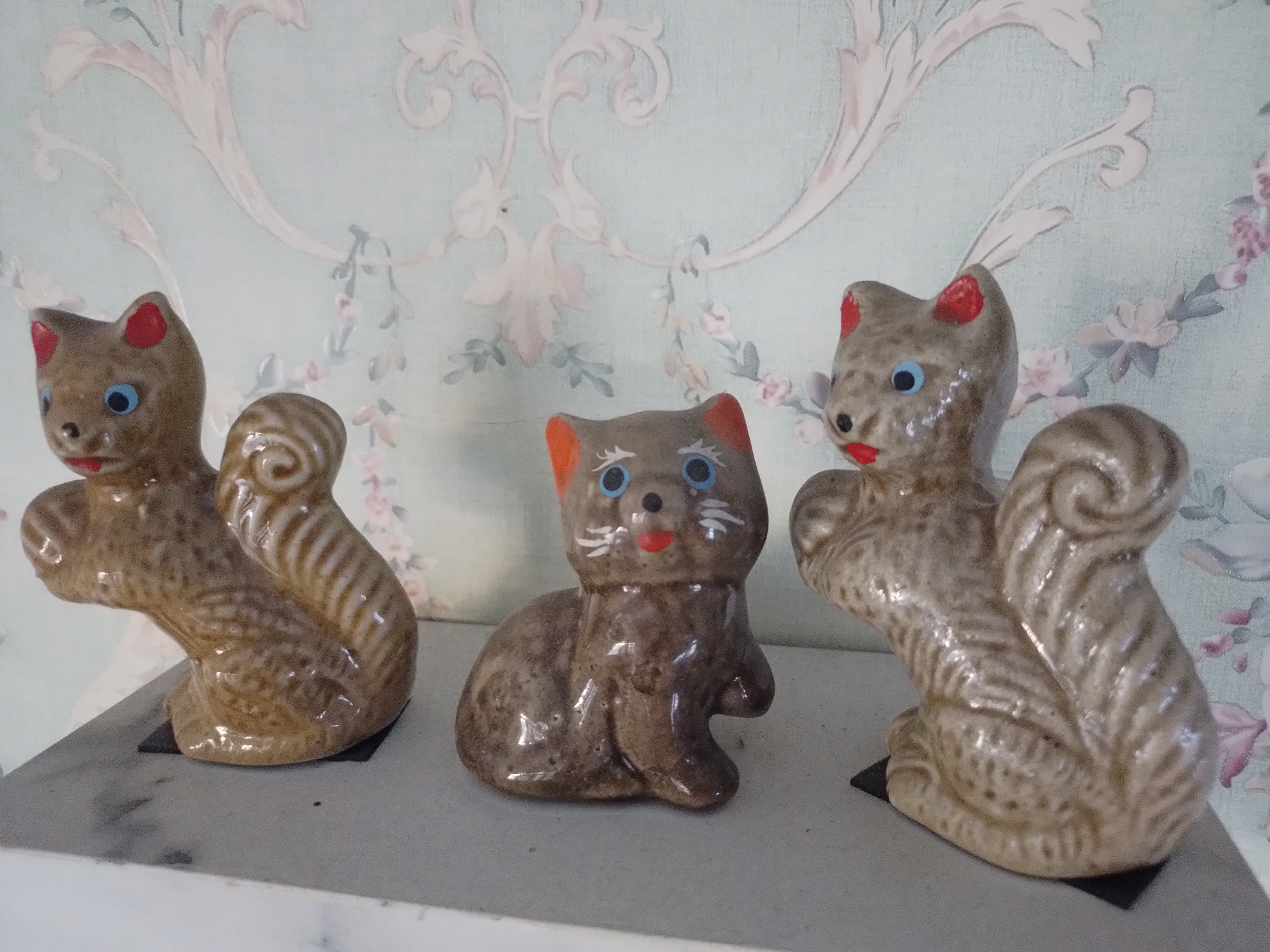Three ceramic figurines of squirrels and one cat are displayed on a shelf against a patterned wallpaper. The two squirrel figurines, one on the left and one on the right, are brown with striped tails and blue eyes. The cat figurine is in the center, brown with orange ears and blue eyes. The wallpaper is light blue with a floral and scroll pattern in pale pink and cream. The shelf is a light gray surface. The image was taken at a residence in Batu, Indonesia.