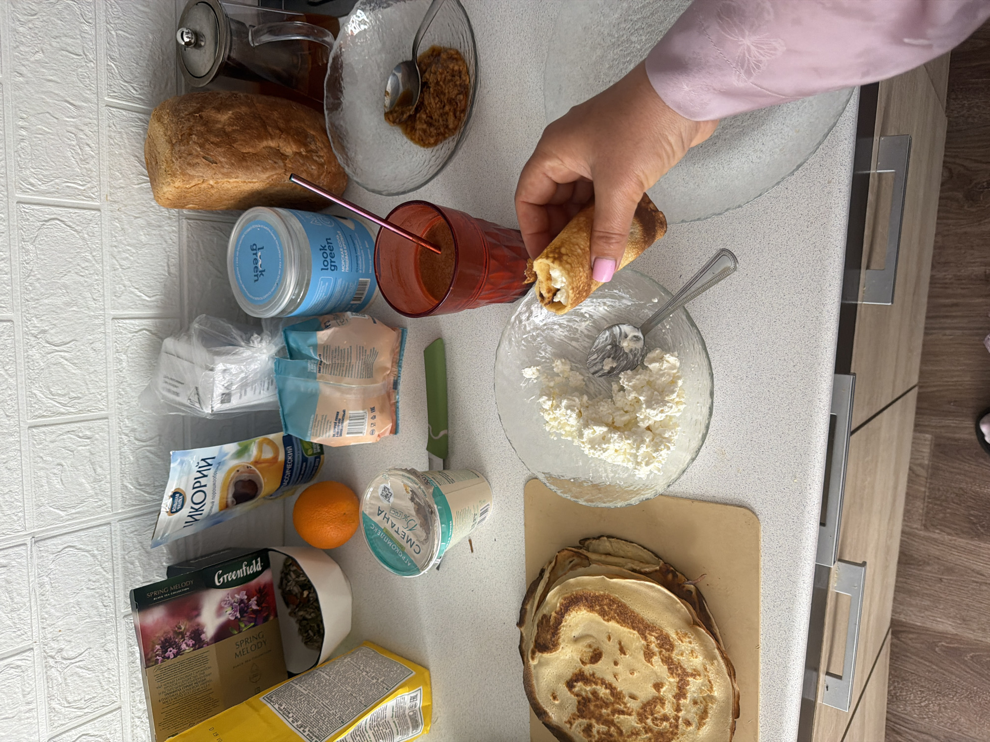 The image shows a top-down view of a kitchen counter cluttered with food items. A hand, presumably belonging to a person wearing a light pink long-sleeved shirt, holds a rolled-up pancake. The pancake is being dipped into a clear glass bowl filled with white cheese (possibly cottage cheese or ricotta).
To the left of the bowl, there is a wooden cutting board with a stack of golden-brown pancakes. Further to the left, there are various food products, including a container of sour cream labeled "Сметана" (Smetana in Russian), an orange, a package of "ИКОРИЙ" (Chicory in Russian), a box of Greenfield "Spring Melody" tea, a yellow box, a clear bag, a jar of "look green" sea salt, a loaf of bread, a clear glass pitcher with a silver lid, and a small clear bowl with a brown condiment. A red glass filled with a dark liquid with a metal straw in it is to the right of the loaf of bread. A green knife lies between the sour cream and the bowl of cottage cheese.
Based on the Russian text on the food products (Сметана, ИКОРИЙ), it is probable that the location is in a Russian-speaking region, potentially in the city of Leningradskaya, Russia. The scene suggests someone is in the process of preparing or eating pancakes.