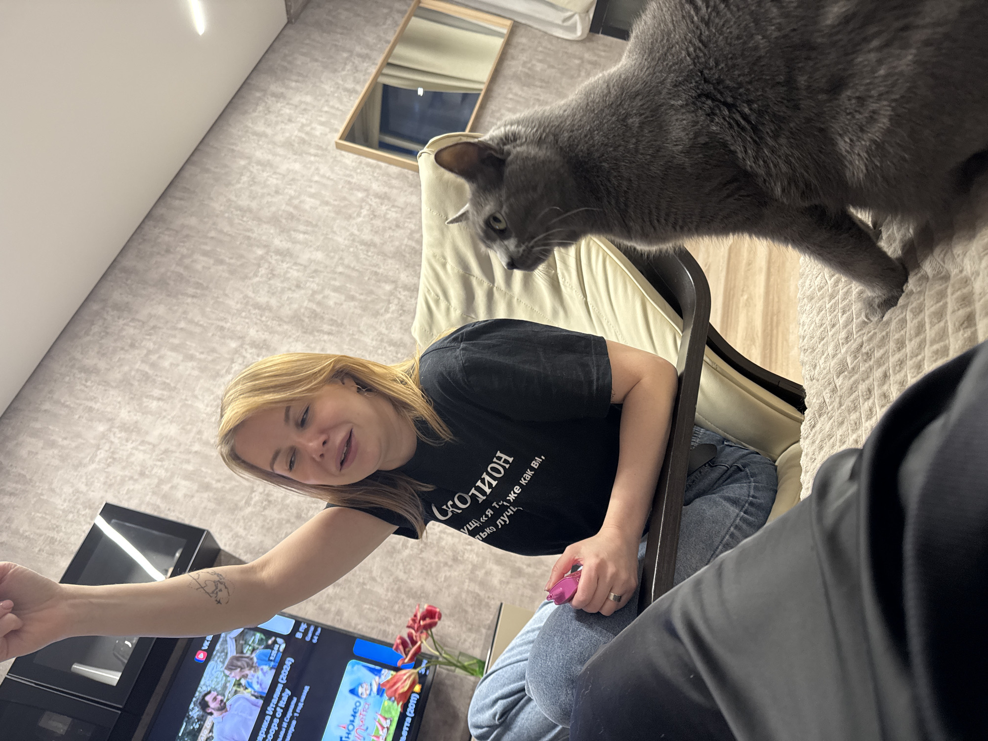 A woman with blonde hair sits in a chair, wearing a black T-shirt with Russian text, jeans, and a silver ring. She holds a small pink object in her right hand. The woman is looking towards the left side of the frame and extending her left arm towards a TV, which shows a program titled "Sartika Italy (2024)". A gray cat is perched on the back of the chair. In the background, there is a mirror, a light-colored wall, and a bouquet of tulips. The scene is indoors.