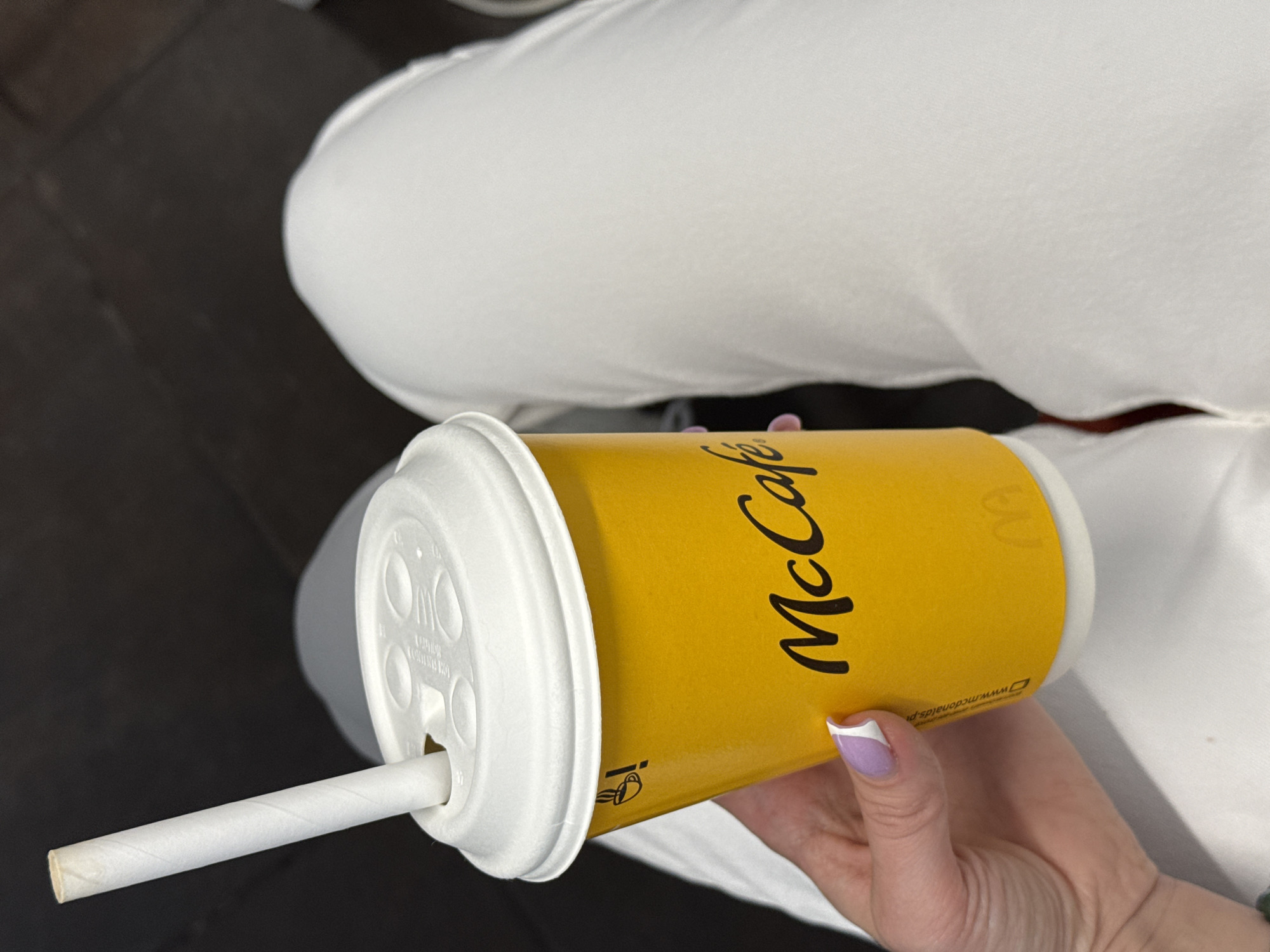 A person's hand, with light purple and white nail polish, holds a yellow McCafe cup. The cup has the McCafe logo printed in black on it. A white paper straw is inserted into the white lid of the cup. The person is wearing white pants, and the background is a dark surface, likely a table or floor. The image appears to be taken indoors, and the lighting suggests it could be daytime. There is visible text on the cup which includes "McCafe" and "www.mcdonalds.pl". The presence of the Polish McDonald's website indicates the location is likely Poland, specifically Łódź as provided in the context.