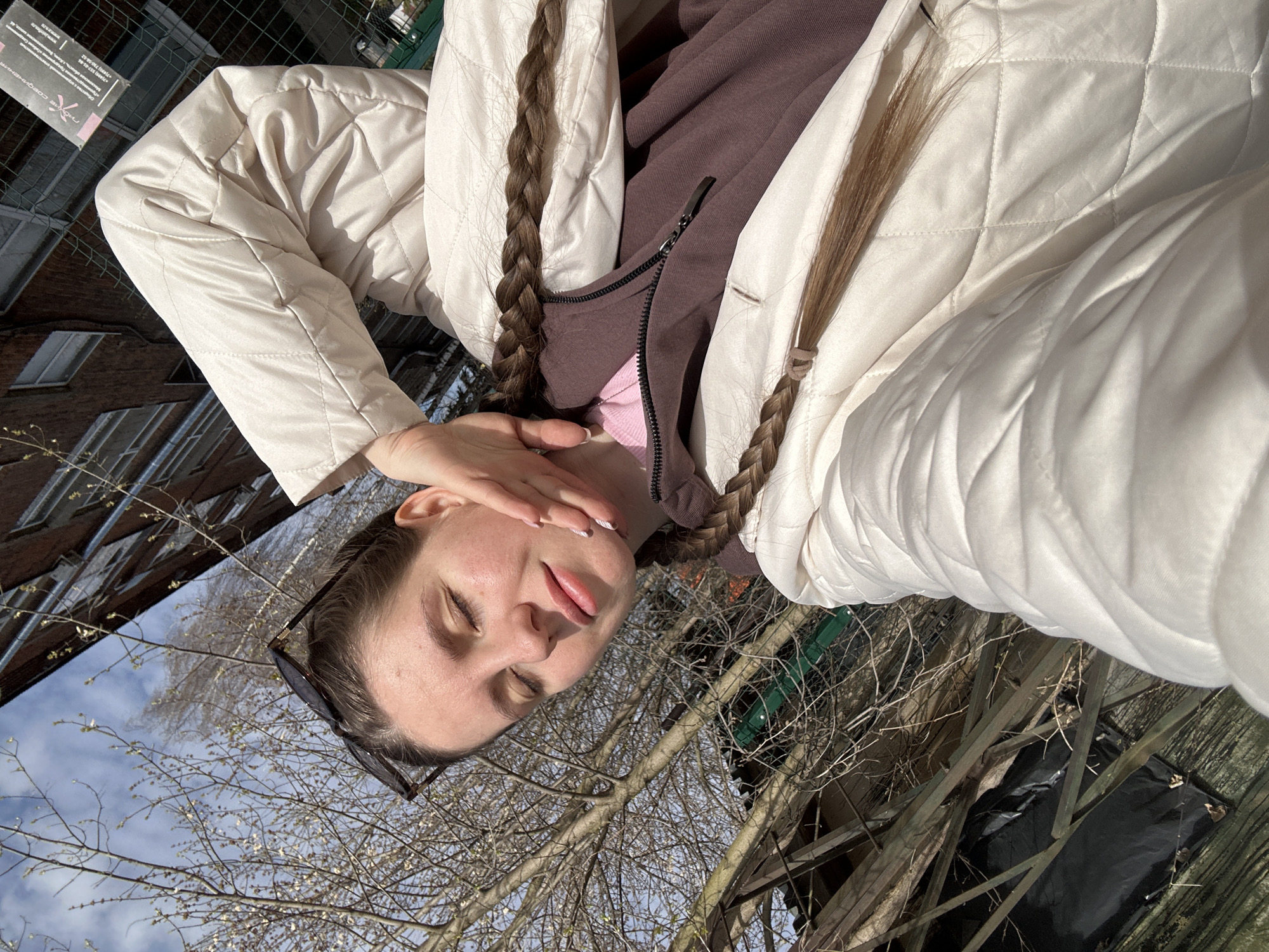 A young woman with long, braided hair is posing outdoors in Klin, Russia. She is wearing sunglasses perched on her head and a light-colored quilted jacket over a brown zipped sweater. She is holding her hand near her face and sticking out her tongue slightly. Behind her are bare trees against a blue sky, a building with brick walls, and a fenced-in area with wood debris and a black tarp.