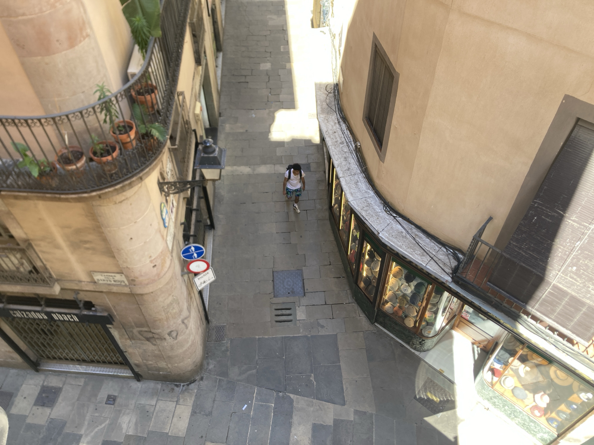 The image captures a high-angle view of a narrow street in Barcelona, Spain. A person is walking down the center of the street. On the left, a building features a balcony with potted plants and a shop with closed metal gate at ground level, "CALZADOS UBACH" is written above the gate. Street signs are visible on the corner of the building. The street is paved with stone, and shadows are cast by the buildings. On the right is a shop with hats displayed in the windows.