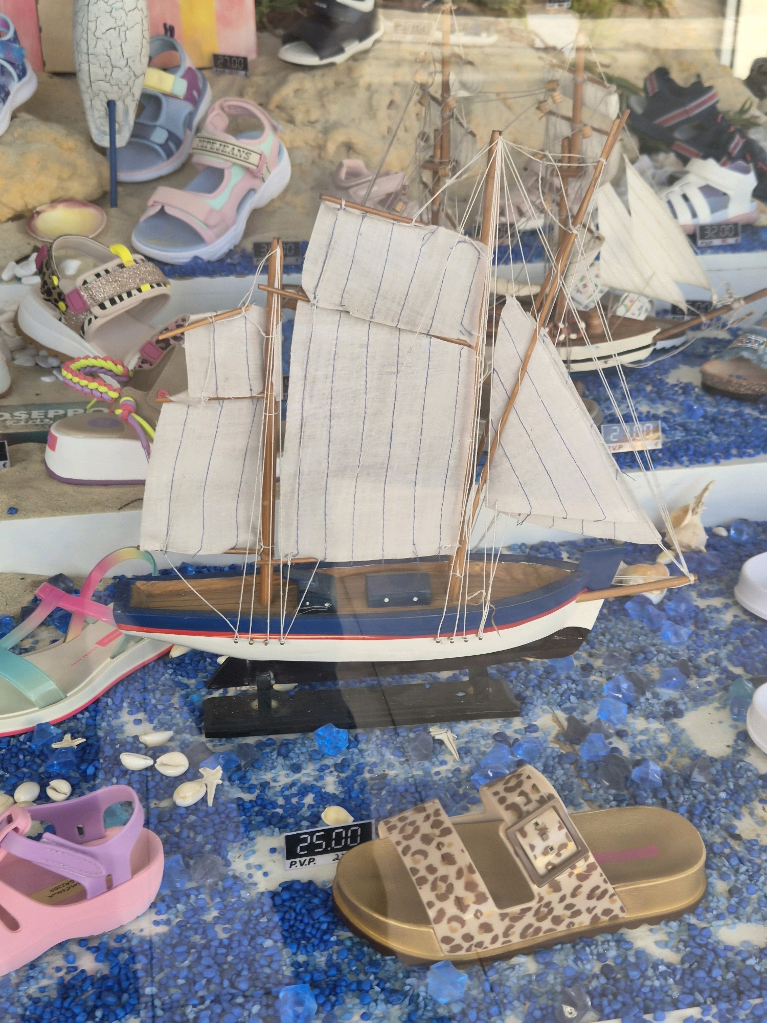 This is a close-up, eye-level, medium shot of a retail store window display in Barcelos, Portugal. The display features a miniature wooden sailboat with white sails, positioned centrally. To the left of the sailboat, several pairs of children's sandals are arranged. These include a pink and blue sandal, a pink sandal with a glittery strap, and a pale pink sandal. In the foreground, a light blue and pink sandal with clear straps is visible. To the right of the sailboat, a single leopard-print slide sandal with a gold sole is on display. The background of the display is filled with small blue and clear decorative stones, along with a few seashells and plastic blue geometric shapes. In the upper portion of the image, glimpses of other retail items, including shoes and clothing, can be seen behind the main display. A price tag with "25.00 P.V.P." is visible in the lower right quadrant. The lighting is consistent and appears to be from artificial sources within the store.