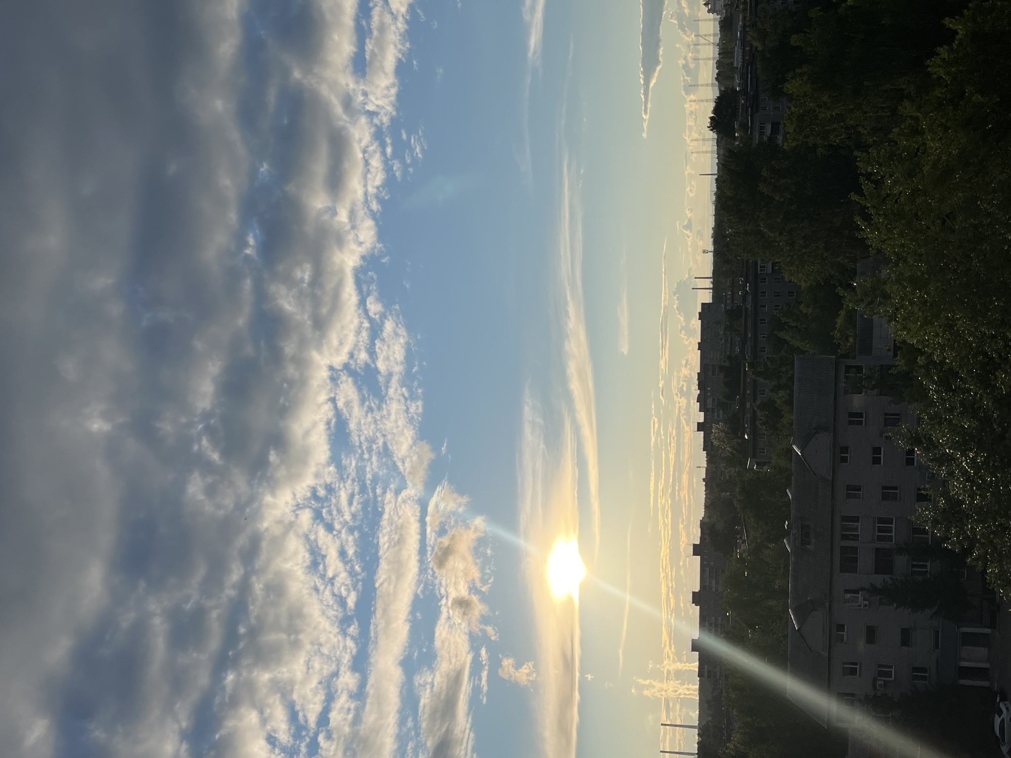 The image depicts a scene in an urban environment, likely a residential area in Unknown, Unknown. The sky dominates the view, with a mix of blue sky and clouds, ranging from thin, wispy cirrus to thicker, more textured cumulus formations. The sun is visible, positioned low in the sky, possibly indicating early morning or late afternoon.
On the right side of the image, there are buildings, seemingly multi-story apartment complexes. Some are partially obscured by trees, suggesting the presence of green spaces within the city. The architectural style appears to be Soviet-era or similar.
Industrial elements, such as smokestacks or power line towers, are visible in the distance, contributing to the urban landscape. The light from the sun casts long shadows, emphasizing the three-dimensional nature of the buildings and trees.