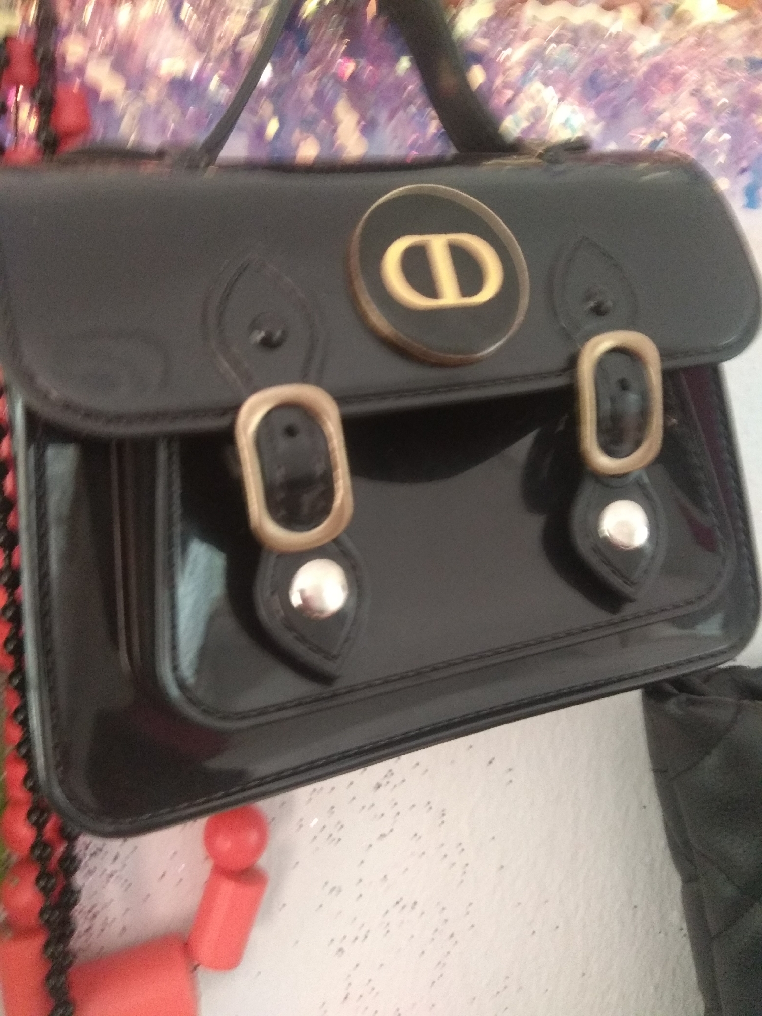 The image shows a close-up of a black patent leather handbag with gold hardware. The bag has a structured, satchel-like design with two front flap closures secured by buckles. A prominent gold "CD" logo, characteristic of the Dior brand, is affixed to the top center of the bag. The background is blurred and appears to be a wall with some sparkly decorations, possibly glitter or confetti, giving a festive or decorative feel. To the left of the bag, a string of red beads is visible, and a bright red toy or decoration with geometric shapes is partially in frame at the bottom. To the right, a quilted black item, possibly another bag or accessory, is partly visible. The lighting suggests an indoor setting, and the overall impression is one of a still-life arrangement showcasing the handbag. There is no discernible activity or event taking place; the image is a static shot of an object. The location context of Jalingo, Nigeria, does not provide any specific visual cues within the image itself. No text is clearly legible on the handbag or any other visible element.