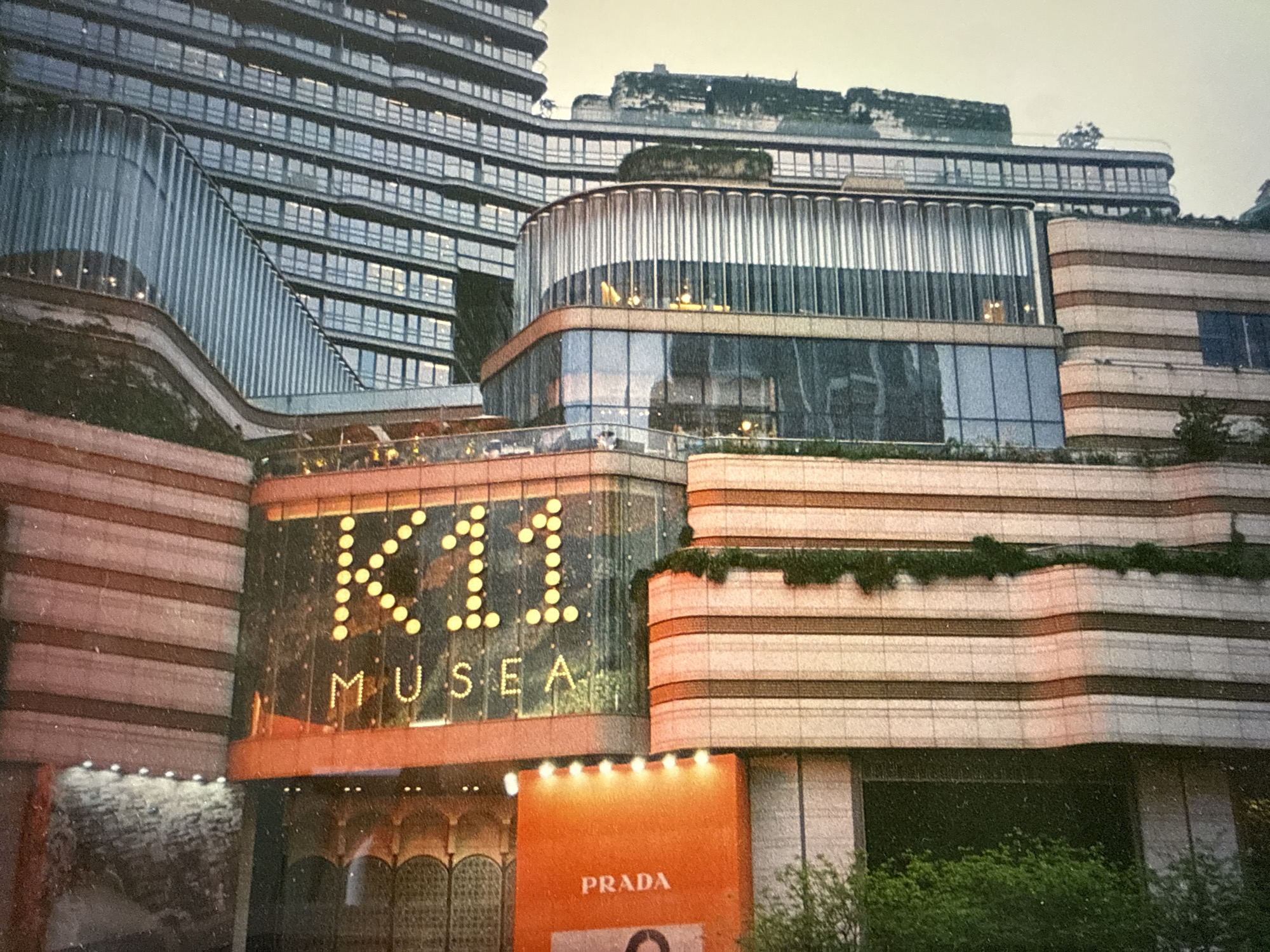 The image depicts the K11 MUSEA, a retail complex. The facade prominently features the illuminated letters "K11" and the word "MUSEA" below. Below that is a shop with a sign reading "PRADA." The architecture of the building involves multiple levels of horizontal striping, varied materials (glass, concrete), and terraced landscaping. The scene does not depict Almaty, Kazakhstan. It is in Hong Kong.