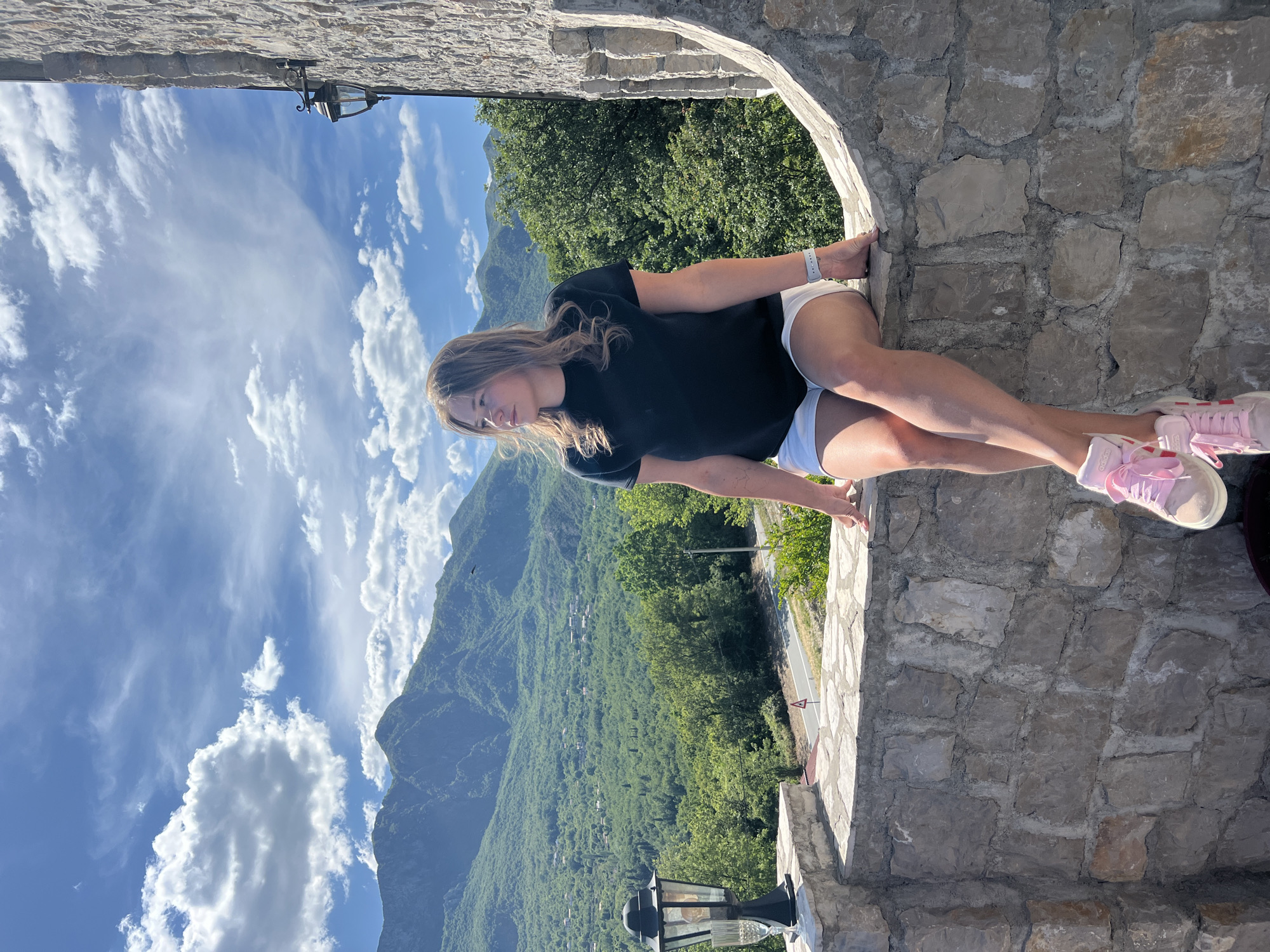 A young woman with light brown hair is seated within a rustic stone archway, gazing out at a panoramic mountain vista. She wears a black t-shirt, white shorts, and light pink sneakers. A grey smartwatch is visible on her left wrist. Her pose is relaxed, with legs crossed at the ankles.
Beyond the archway, a vast, verdant mountain range stretches under a bright blue sky dotted with white, wispy clouds. The slopes are densely covered with lush green trees. In the valley below, a winding road can be seen, along with scattered buildings, suggesting a small village or settlement nestled amidst the natural landscape. A black, classic-style street lamp with a glass cover is affixed to the stone wall in the lower foreground.
The scene is brightly lit by natural sunlight, indicating a clear, sunny day, likely mid-morning or afternoon. The stone archway is constructed from rough-hewn, irregular grey stones cemented with visible mortar. The woman's sneakers clearly show the brand "adidas." A small, triangular traffic sign with a red border is partially visible near the road. The setting provides a scenic overlook, capturing the natural beauty of Sotonići, Montenegro, with its mountainous terrain and clear skies.