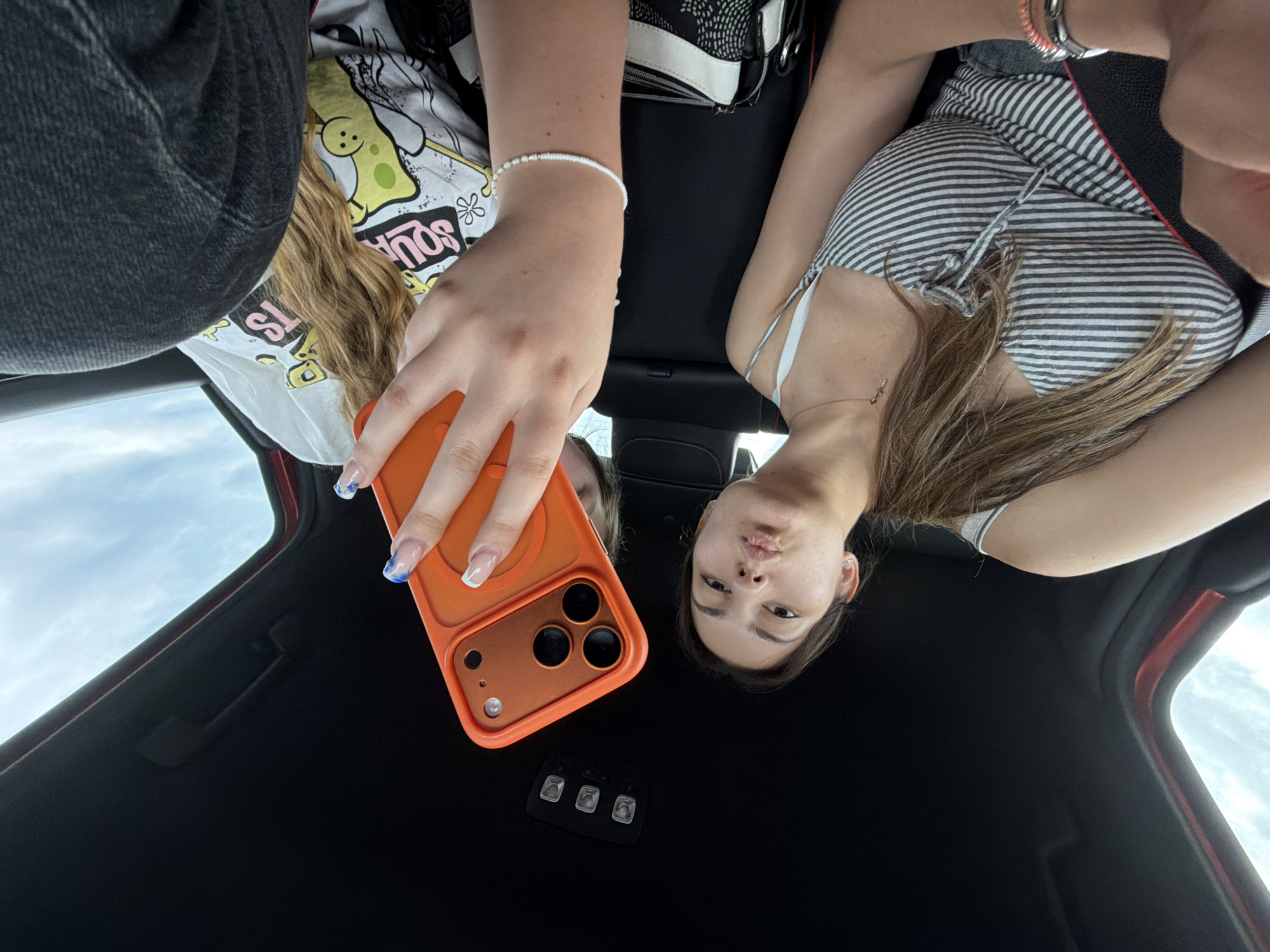 The image shows two people in a car, likely taking a selfie. The camera is positioned looking upward toward the car's roof. The person on the left is wearing a Spongebob Squarepants t-shirt and jeans. They are holding an orange iPhone. The person on the right is wearing a striped top. The car's interior is primarily black with red trim. The environment outside appears to be a cloudy sky. This photo was not taken in Koniovo, Bulgaria.