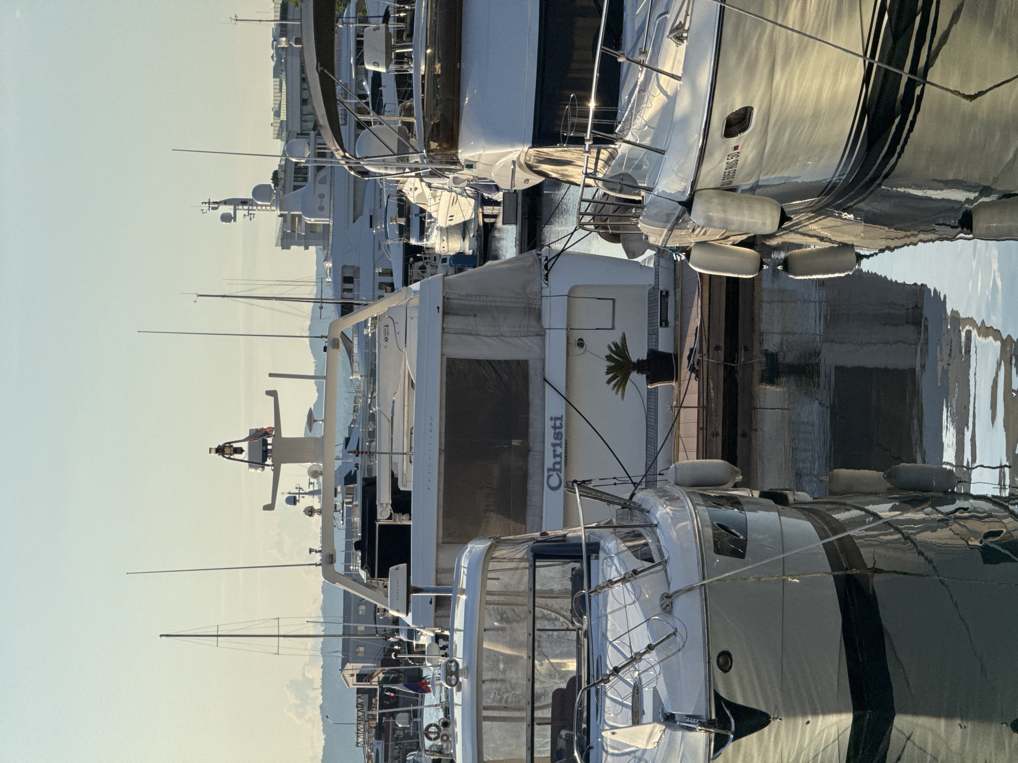 The image displays a marina filled with docked yachts and boats. The scene is set in Sochi, Russia, under a clear, bright sky, suggesting it is daytime, possibly late afternoon due to the golden light reflecting off the water and the polished surfaces of the boats. The water is calm, mirroring the vessels and the sky, creating a tranquil atmosphere.
Several yachts of varying sizes are visible, their sleek designs and white hulls prominent against the blue of the water and sky. Some boats have dark accents, adding to their modern aesthetic. Fenders are strategically placed along the sides of the larger yachts to protect them from impact. One prominent yacht has the name "Christi" inscribed on its side in elegant lettering. Another yacht has text that appears to read "RODEO KING 50".
The marina setting is evident with the presence of docks and mooring lines. In the background, buildings and a hazy mountain range can be seen, hinting at the coastal urban environment of Sochi. The overall impression is one of luxury and leisure, with the boats appearing well-maintained and ready for use. The lighting suggests a pleasant day for boating.