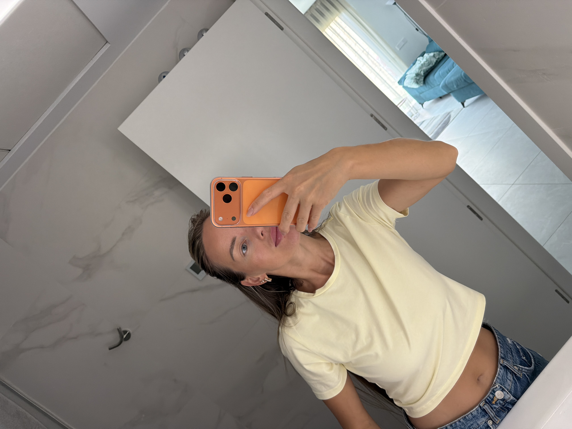 A woman stands in front of a mirror in a bathroom, taking a selfie with her orange-cased smartphone. She is wearing a light yellow t-shirt and blue jeans, with her midriff exposed. Her long blonde hair is pulled back, and she is wearing small hoop earrings. The bathroom features marble-patterned walls and white cabinetry. In the reflection, a turquoise sofa with a patterned cushion is visible in what appears to be a living area. The scene is set in Finestrat, Spain.