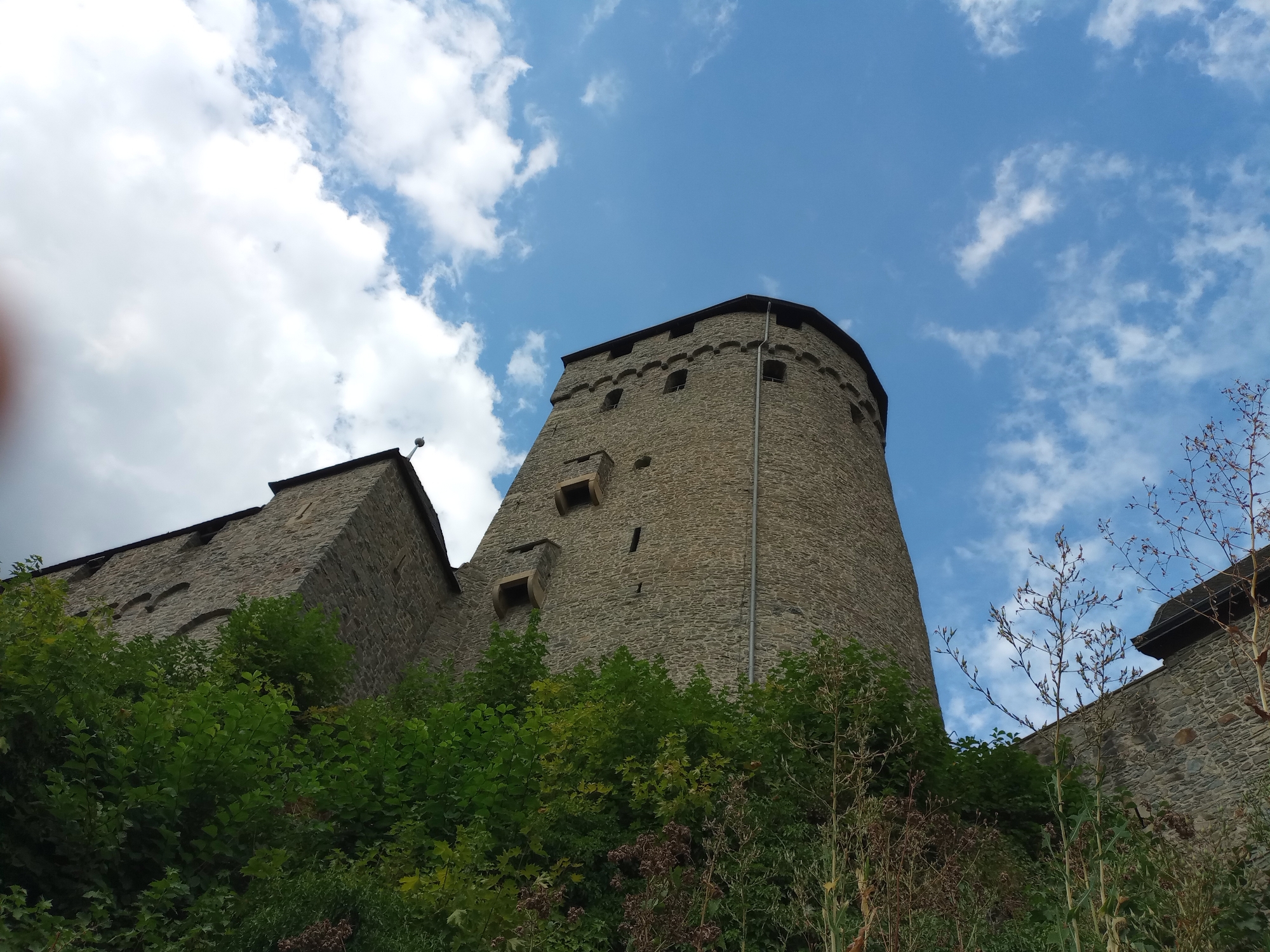 The image shows a view from below of a stone castle on a hill, with lush green foliage in the foreground. The castle has a tall, round tower with several small windows and a crenellated top. To the left of the round tower is a section of the castle wall with a pitched roof. The sky above is blue with scattered white clouds. The foliage is dense and appears to be trees and bushes. The overall impression is that of an ancient fortress set against a natural landscape. This is a view of Altena Castle, located in Altena, Germany.