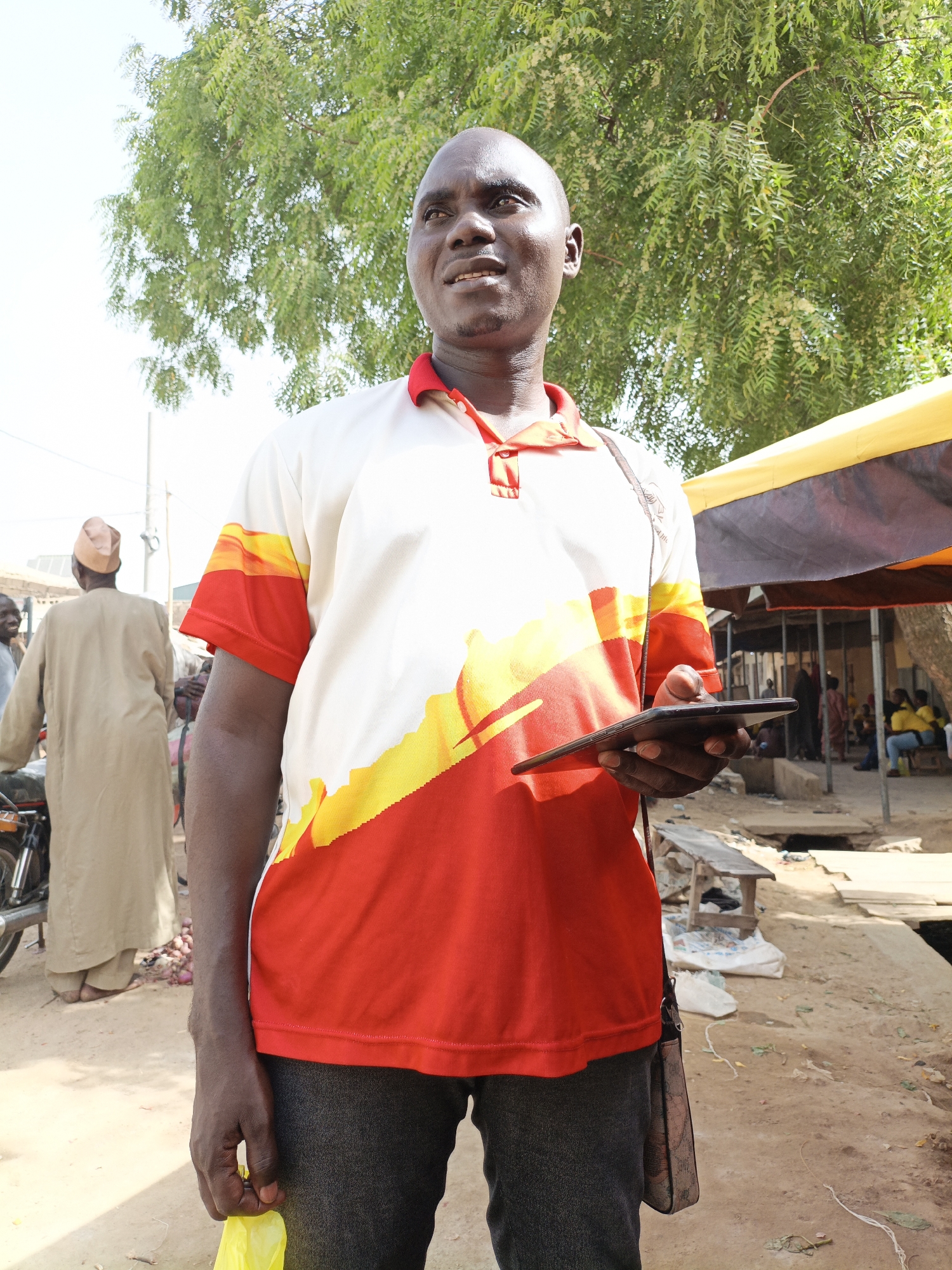 In Kawaji, Nigeria, a bald, dark-skinned man in a white, red, and yellow shirt and grey pants stands outside holding a tablet, looking to his left. He has a bag slung over his shoulder and a yellow plastic bag in his right hand. Behind him, there is a tree, a building with a brown and yellow awning, and other people. A man in a tan robe stands by a motorcycle to the left. The ground is dirt with litter scattered around.