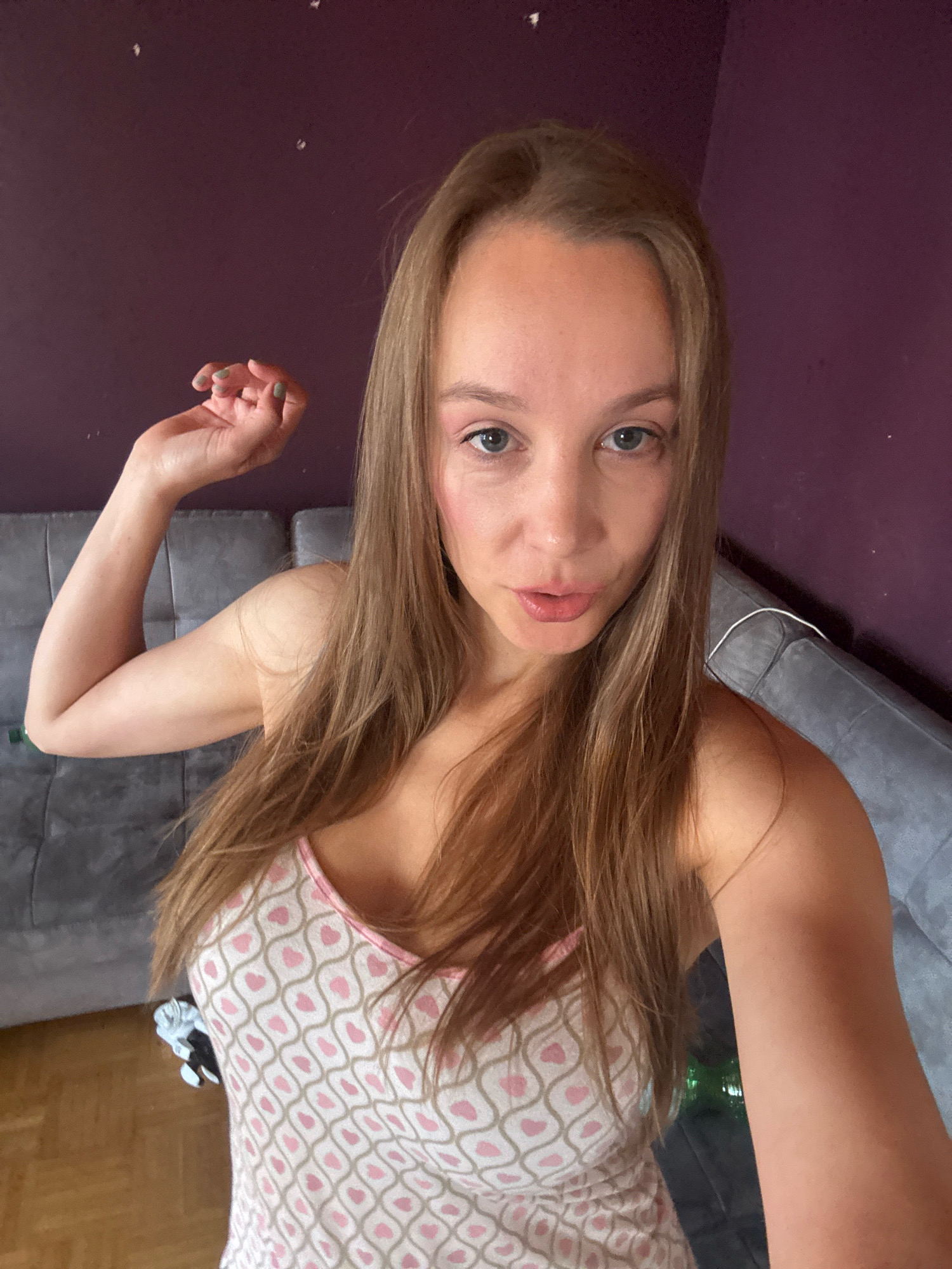 The image shows a woman with long, light brown hair, wearing a tank top with a pink and beige heart pattern. She is indoors, in what appears to be a bedroom, with a purple wall behind her and a grey sofa visible. The woman is taking a selfie, with her left arm raised and hand near her head. She is looking at the camera with a slightly puckered expression. The lighting suggests it is daytime, possibly with natural light coming from a window. There are no discernible landmarks or features that would indicate a specific location within Überherrn, Germany.