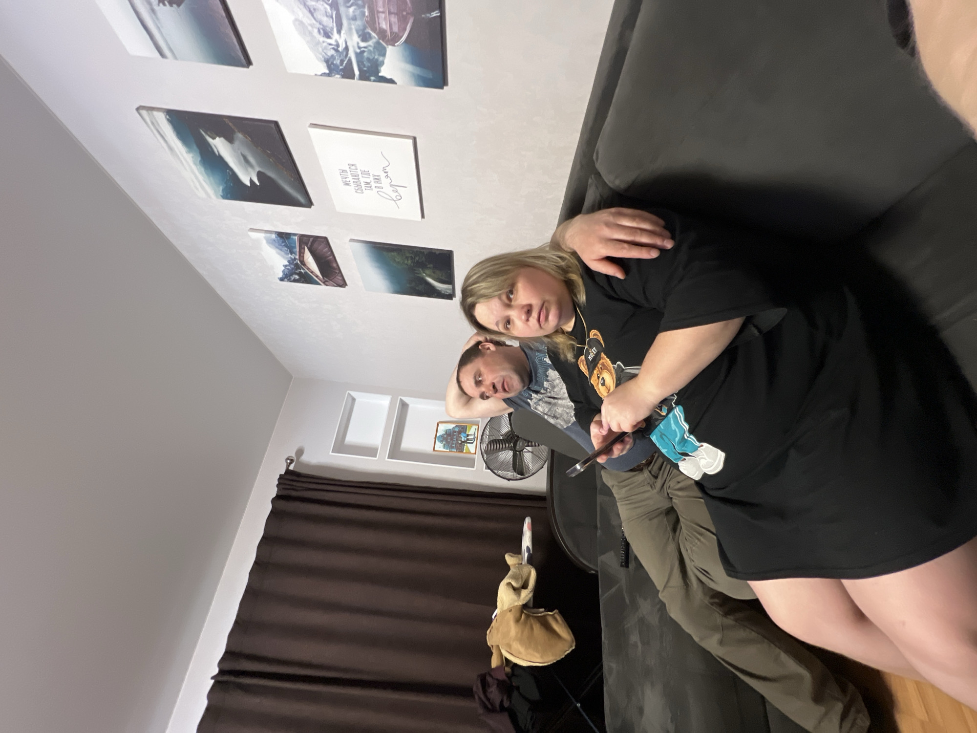 This image captures two individuals relaxing indoors, likely in a living room in Perm, Russia. A woman and a man are seated closely on a dark grey sofa. The woman, with blonde hair, wears a black t-shirt featuring a cartoon character, possibly a bear. She holds a smartphone or similar device in her hands, and her left arm rests around the man's shoulder. The man has dark hair and a beard, dressed in a grey/blue patterned t-shirt and olive green trousers. He leans back against the sofa, with his right hand behind his head, appearing relaxed.
The room features light-colored walls adorned with several framed pictures or canvases, depicting natural scenes like mountains and water. Below these, there are two white square shelves. One of the canvases displays Cyrillic text, which translates to "DREAMS COME TRUE WHERE they are believed in." A dark brown curtain covers a window on the left side of the frame. A standing fan is visible behind the man, and a bag hangs from a stand or hook further back. The overall atmosphere appears casual and comfortable, with both individuals displaying neutral and relaxed expressions. The lighting suggests it could be daytime or evening, with indoor illumination.
