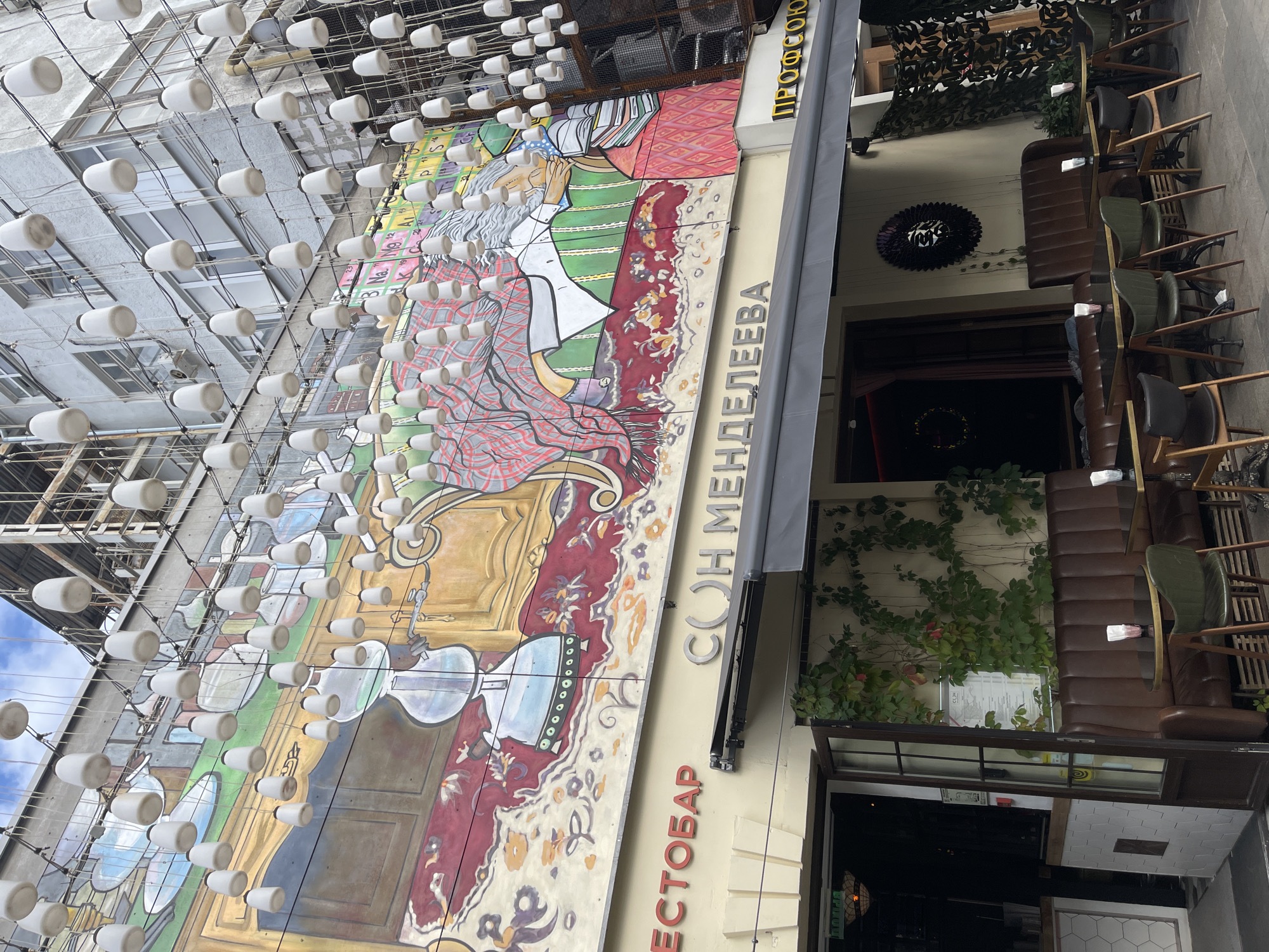 The image captures the facade of a restaurant named "СОН МЕНДЕЛЕЕВА" (Mendeleev's Dream) in Moscow, Russia. Above the entrance, a vibrant mural depicts a seated figure, possibly representing Dmitri Mendeleev, surrounded by elements related to chemistry, such as the periodic table. The mural is further enhanced by numerous white, bell-shaped lanterns suspended by wires, creating a canopy-like effect.
The restaurant features an inviting outdoor seating area with dark brown leather banquettes and small, dark-topped tables. Various chairs, some in green and others in wood and black, are arranged around the tables. Green ivy climbs the wall behind the seating area. The entrance to the restaurant is a glass-paneled door, partially open, revealing a glimpse of the interior. To the left of the main entrance, a sign with red lettering reads "РЕСТОБАР" (Restobar). To the right, yellow lettering spells out "ПРОФСОЮЗ" (Profsoyuz), possibly indicating an associated establishment or a nearby location. The overall scene suggests a stylish and artistic establishment in a bustling urban setting.