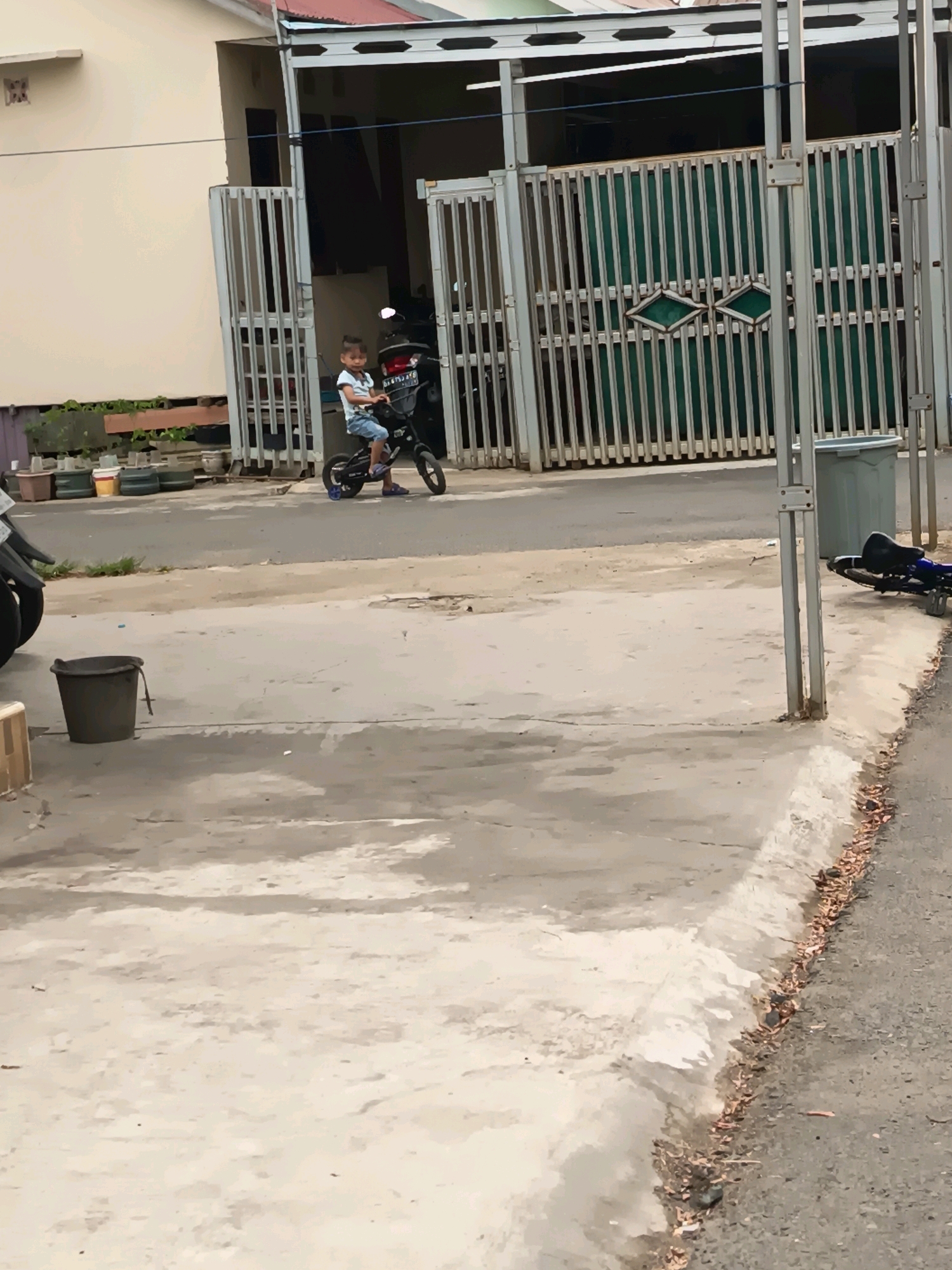 The image depicts an outdoor scene, likely in a residential area of Banjarmasin, Indonesia, given the style of the house and gate. In the foreground, a concrete walkway and road are visible. To the left, a black bucket sits on the walkway. Further back, a young boy is riding a small black electric motorcycle. He is wearing a light-colored t-shirt and blue shorts. The motorcycle has a license plate that appears to be from Indonesia. Behind the boy is a residential building with a white facade and a metal gate. The gate is closed and has a green panel behind it. The overall environment suggests a warm, possibly sunny day.
