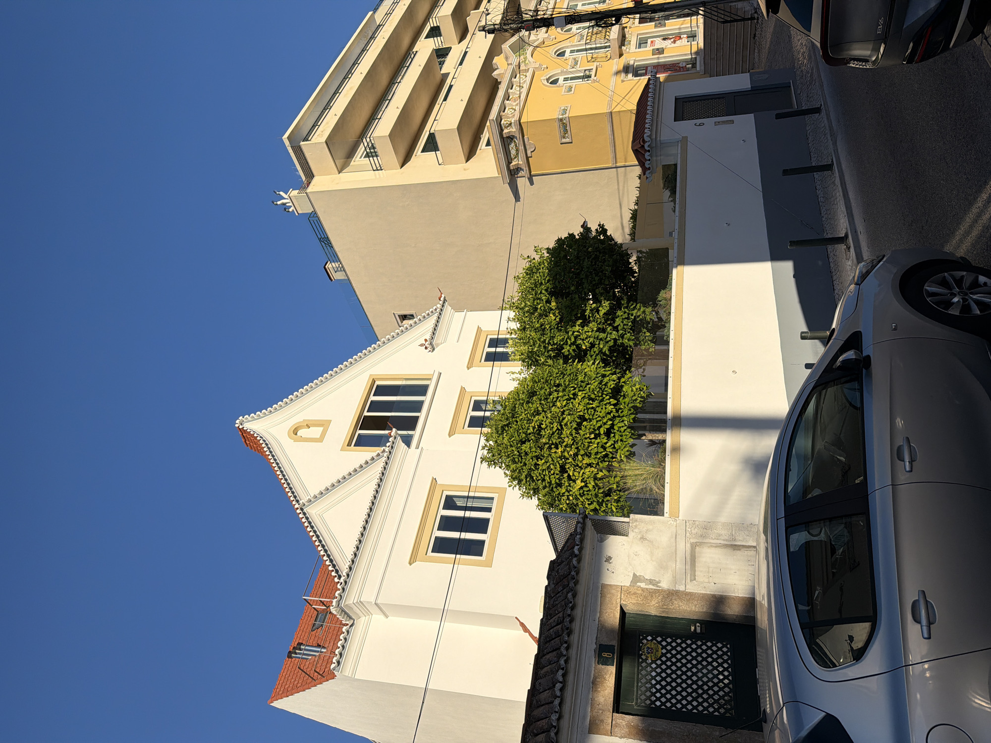 This image depicts a street scene in Cascais, Portugal, with a clear blue sky suggesting it's daytime and pleasant weather. The perspective is from a low angle, looking upwards at a cluster of buildings and a parked car.
Dominating the background is a tall, modern apartment building with a light beige facade and balconies. Adjacent to it is a narrower, yellow building with ornate architectural details, including arched windows and decorative elements on its facade. In the foreground, a charming, white two-story house with a red-tiled roof and intricate white trim around its gables and eaves stands out. It has several windows with yellow surrounds. A lush green tree grows beside this house, adding a touch of nature to the urban setting.
A silver car is parked on the right side of the street, partially obscuring a darker car further down. The street itself appears to be paved with cobblestones. Visible on the white house is a dark green door with a patterned grate, and a small number "8" above it. There are also power lines running across the scene, a common sight in urban environments. The sunlight casts distinct shadows, indicating the sun is at an angle, typical of late afternoon or early morning. No people are visible in the image.