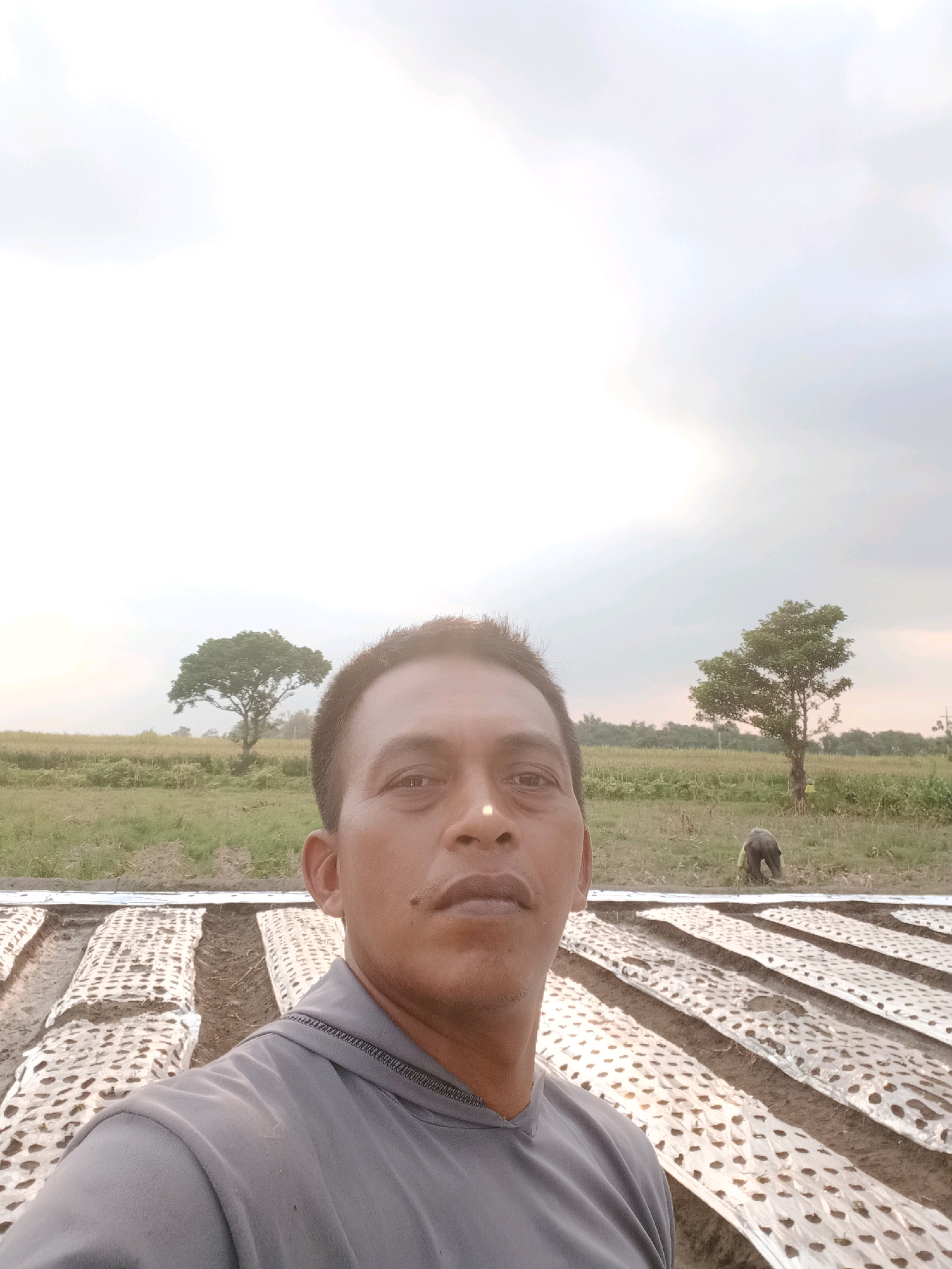 A man, with dark skin and short, dark hair, takes a selfie in an outdoor field. He is wearing a gray hooded sweatshirt. The man's face is close to the camera, and the background shows a field with rows of covered soil. In the distance, there are trees and a person working. The sky is overcast and light.
