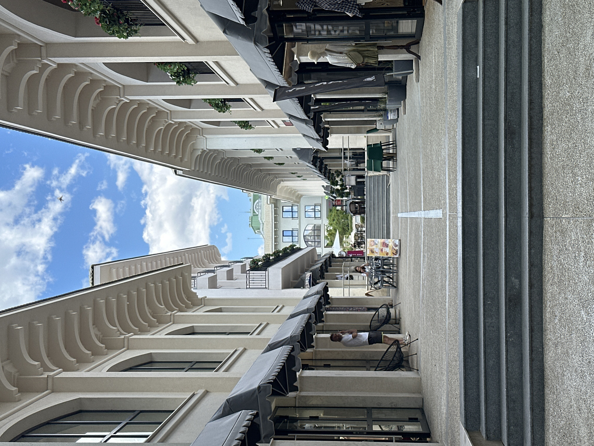 The image depicts a perspective shot of a pedestrian area in Varna, Bulgaria. On either side are buildings with arched colonnades, awnings, and planter boxes on the upper levels. People are seated at tables in the pedestrian walkway, indicating outdoor cafe or restaurant seating. A man in a white shirt and dark shorts is seated on a chair near the bottom of the image. In the center are stairs leading upwards. The sky is visible between the buildings with some clouds and a single bird. The style is modern European with architectural details reminiscent of Mediterranean design.