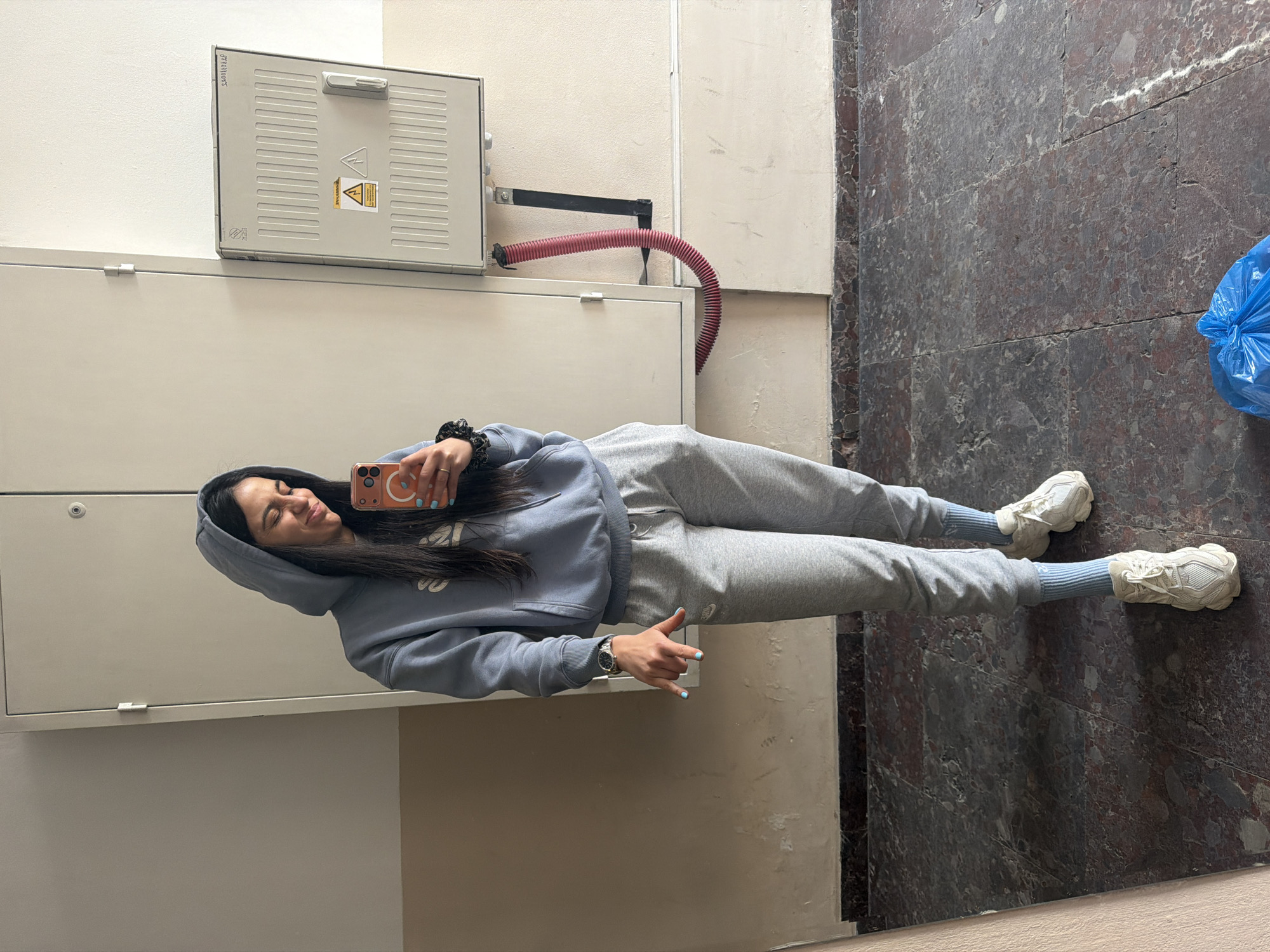 A young woman is posing for a selfie in a corridor. She is wearing a light blue hoodie, grey sweatpants, striped socks, and white sneakers. She holds an orange smartphone in her right hand, while making a "rock on" gesture with her left. Her expression is relaxed. The backdrop features a wall with a beige lower section and a grey upper section, where a large grey electrical box is mounted. A red corrugated pipe extends from the box. The floor is made of dark brown marble tiles. A blue plastic bag is visible on the floor. The location is possibly an interior space in Sofia, Bulgaria.