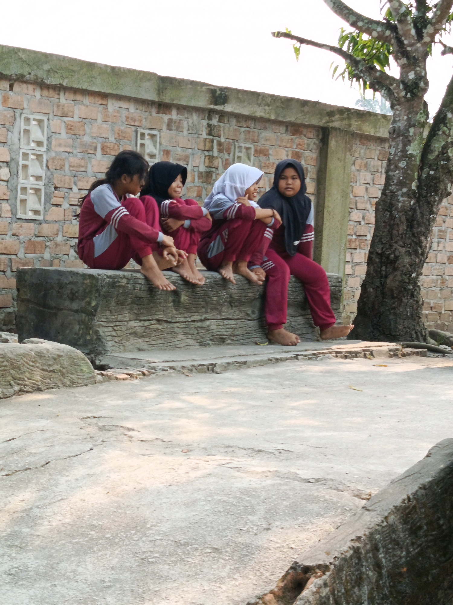 Four young girls, dressed in maroon and gray school uniforms with headscarves, are seated on a rough, weathered stone bench outside a brick building. The girls are positioned from left to right, with the leftmost girl looking away from the camera, the second from the left looking forward and smiling, the third from the left looking slightly to her right, and the rightmost girl looking forward. The building behind them has a rough, textured brick wall with some rectangular openings and is topped by a concrete ledge. To the right of the building, a large tree with dark, textured bark and sparse green leaves stands. The ground in front of the bench is a cracked concrete surface, and a large, irregular rock is visible to the left. The scene appears to be an outdoor area in Karya Dadi, Indonesia, possibly a school or community space, during daylight hours with soft lighting.