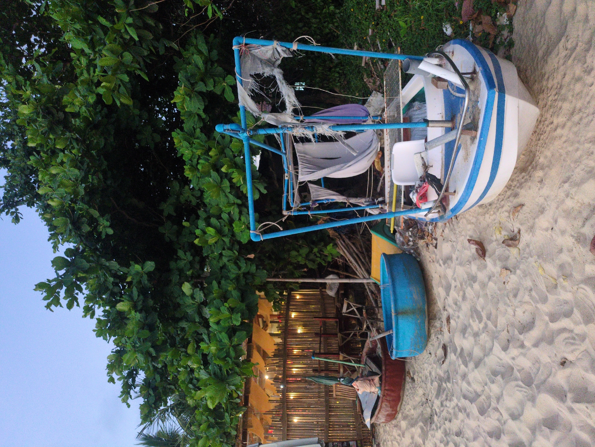 This image, possibly taken in Phú Quốc, Vietnam, shows two boats resting on a sandy beach adjacent to a tree line and a structure with lights. The first boat, with white and blue stripes, has a blue frame structure erected above it, with tattered fabric draped over the frame. A white plastic chair sits inside the boat. The second boat, painted red and blue, lies next to the first. Behind the boats, there's a fence-like structure adorned with strings of lights. The environment is a beach, evidenced by the sand, vegetation, and possibly the location within a coastal city. No people are visible in the image.