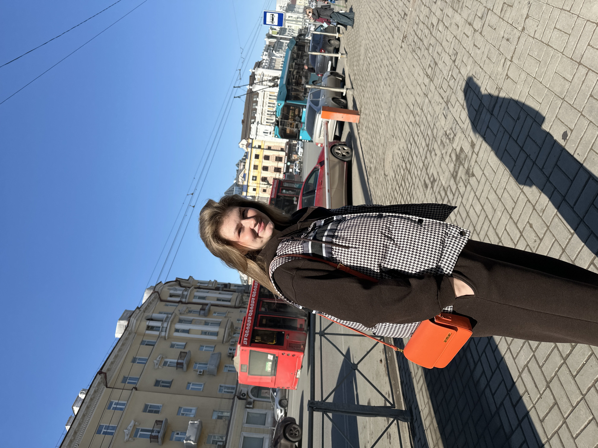 The image displays a young adult female standing on a paved sidewalk under a clear blue sky on Ulitsa Pushkina, Kazan, Russia. She is positioned mid-frame, facing the viewer with a subtle smile. She wears a black long-sleeved top, a black-and-white houndstooth pattern sleeveless puffer vest, black wide-leg trousers, and carries an orange shoulder bag over her right side. Her right hand is inserted into her trouser pocket.
To the subject's left, a grey metal pedestrian barrier runs parallel to the sidewalk, separating it from a multi-lane street. The street contains various vehicles, including a red bus, a teal trolleybus, a red sedan, a silver sedan, and other cars, indicating active traffic. Overhead electrical wires are visible against the sky. On the opposite side of the street, a prominent multi-story, light yellow building with multiple balconies and classical architectural details is visible, along with other urban structures. A white and blue bus stop sign is present on the far right sidewalk. Strong sunlight casts distinct shadows, including a partial human silhouette, onto the light grey interlocking paver sidewalk in the foreground.
