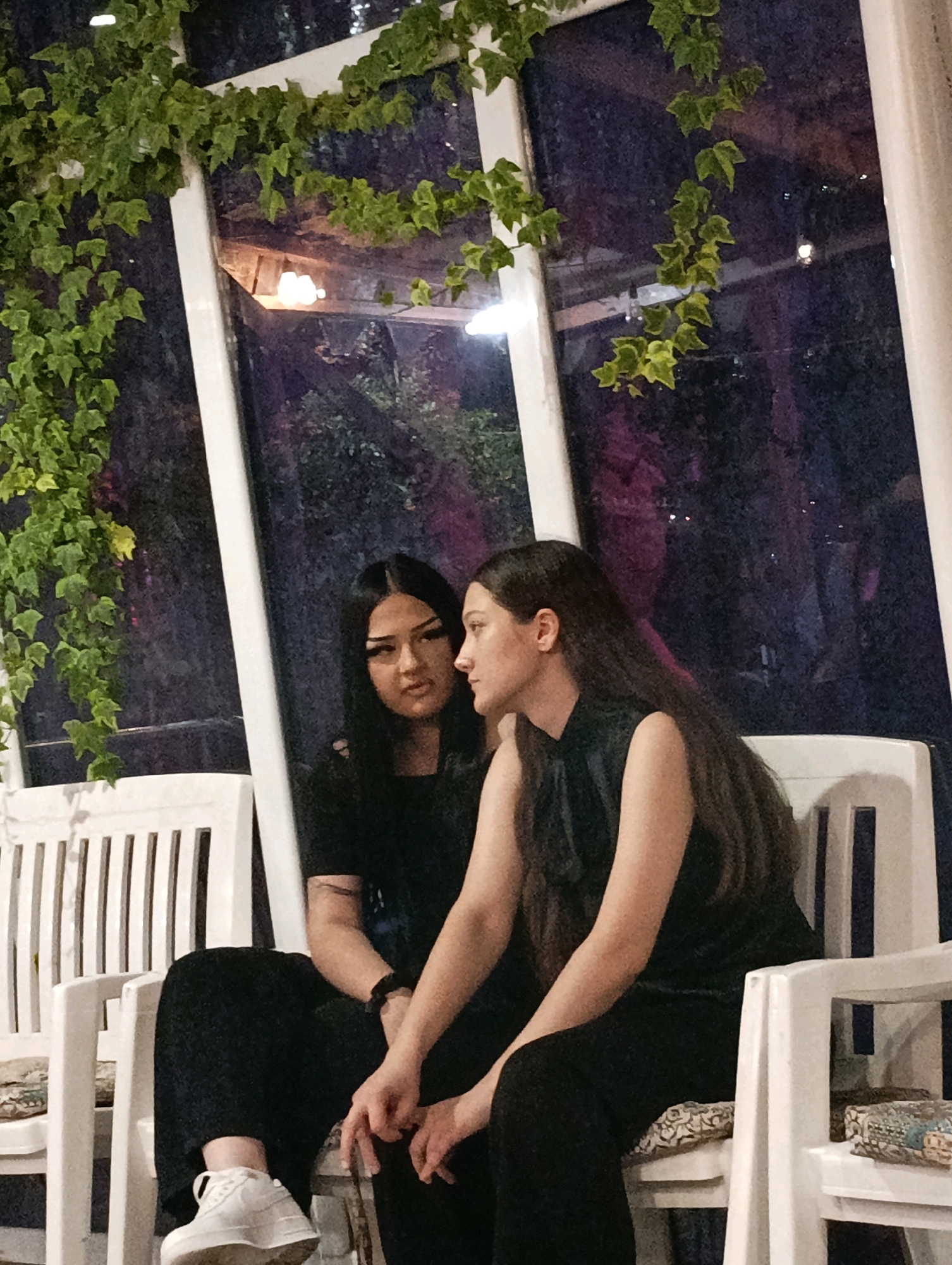 Two young women are seated on white plastic chairs on a patio or outdoor seating area at night. The woman on the left is facing forward with her head turned slightly to her left, looking towards the camera with a neutral expression. She has long, dark hair and is wearing a black top and black pants. Her left hand rests on her lap, and her right hand is near her left thigh. She is wearing white sneakers. The woman on the right is facing left, with her profile visible. She has long, dark hair and is wearing a dark, sleeveless top and dark pants. Her left arm is resting on her lap, and her right arm is bent with her hand near her face. Both women are seated in front of a large window or glass wall that reflects the outdoor environment. Green ivy vines are draped across the top of the window frame and hang down along the sides, creating a naturalistic ambiance. The background outside the window is dark, with some lights and greenery visible, suggesting an outdoor setting at night, possibly in a restaurant or garden. The chairs have slatted backs and seats, and there are cushions on the seats. The overall lighting is dim, with some artificial lights illuminating the scene. The location is identified as Korzhevo, Moldova.