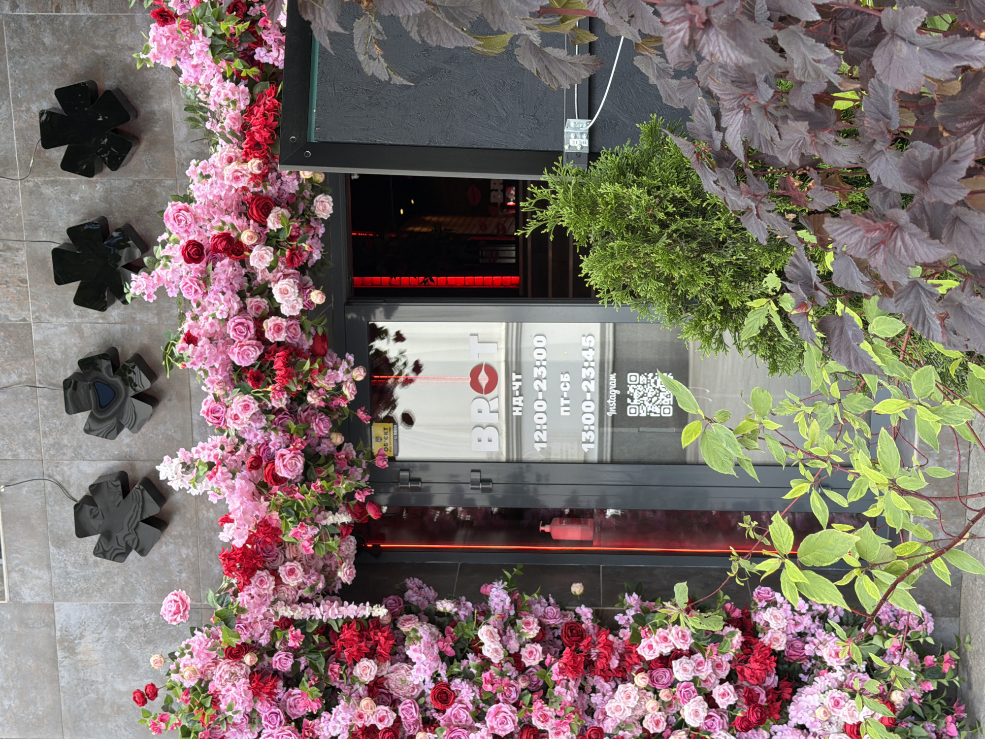 The image shows the entrance to a business called "BROT" in Zhytomyr, Ukraine. The entrance is heavily decorated with a floral arrangement of pink and red roses and other flowers. A glass door leads into a dark interior with red lighting. Above the door, there are three black, abstract, cross-shaped light fixtures mounted on a tiled wall. To the right of the door, there is a black textured panel with some wiring and a metal bracket attached. In front of the entrance, there are several plants, including a small green evergreen and a plant with dark purple leaves. The scene appears to be outdoors, possibly on a street. The time of day is not discernible from the image. There are no people visible in the scene.