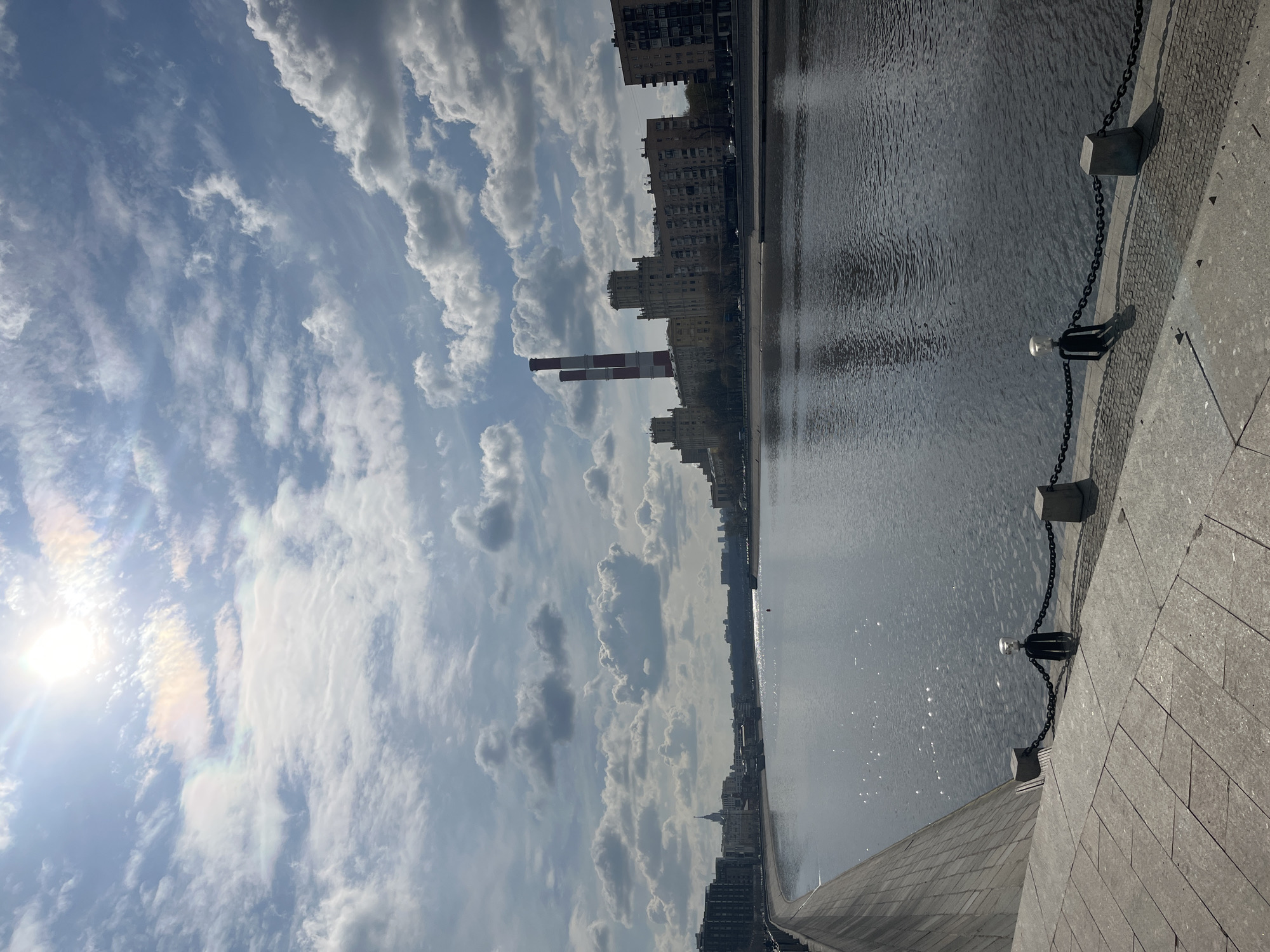 The image depicts a waterside view in Moscow, Russia, characterized by a mix of industrial and urban elements under a partly cloudy sky. In the mid-ground, tall buildings line the waterside, interspersed with what appears to be industrial chimneys, suggesting the presence of a power plant or similar facility. The water reflects the sky and buildings, creating a shimmering effect. A stone embankment with chains and posts runs along the edge of the waterway in the foreground. The sun, visible in the sky, contributes to the overall brightness of the scene.