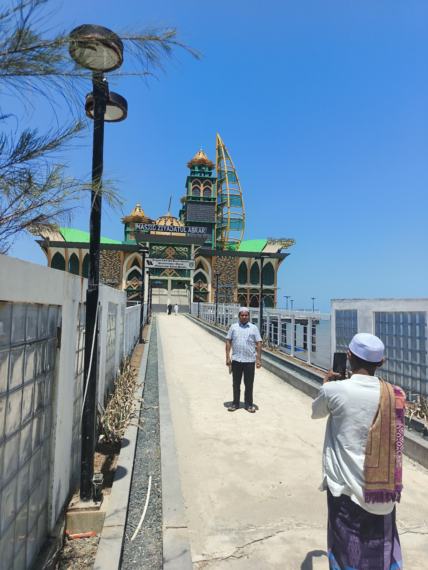 In the city of Beringin, Indonesia, a man in a white skullcap and a purple sarong is captured from behind, holding a smartphone to photograph another man standing further down a paved walkway. The man being photographed, dressed in a checkered shirt and dark pants, faces away from the camera, looking towards a large, ornate building. The building, identified by a sign as "MASJID ZIYADATUL ABRAR," features green and gold architectural elements, reminiscent of Islamic design. To the left of the walkway, a white fence with glass block sections lines the path. A lamppost stands prominently on the left side of the frame, partially obscured by branches from an unseen tree. The environment appears to be outdoors, under a clear blue sky, with the sea visible in the background, suggesting a coastal location. The overall scene depicts a moment of tourism or religious site visit.