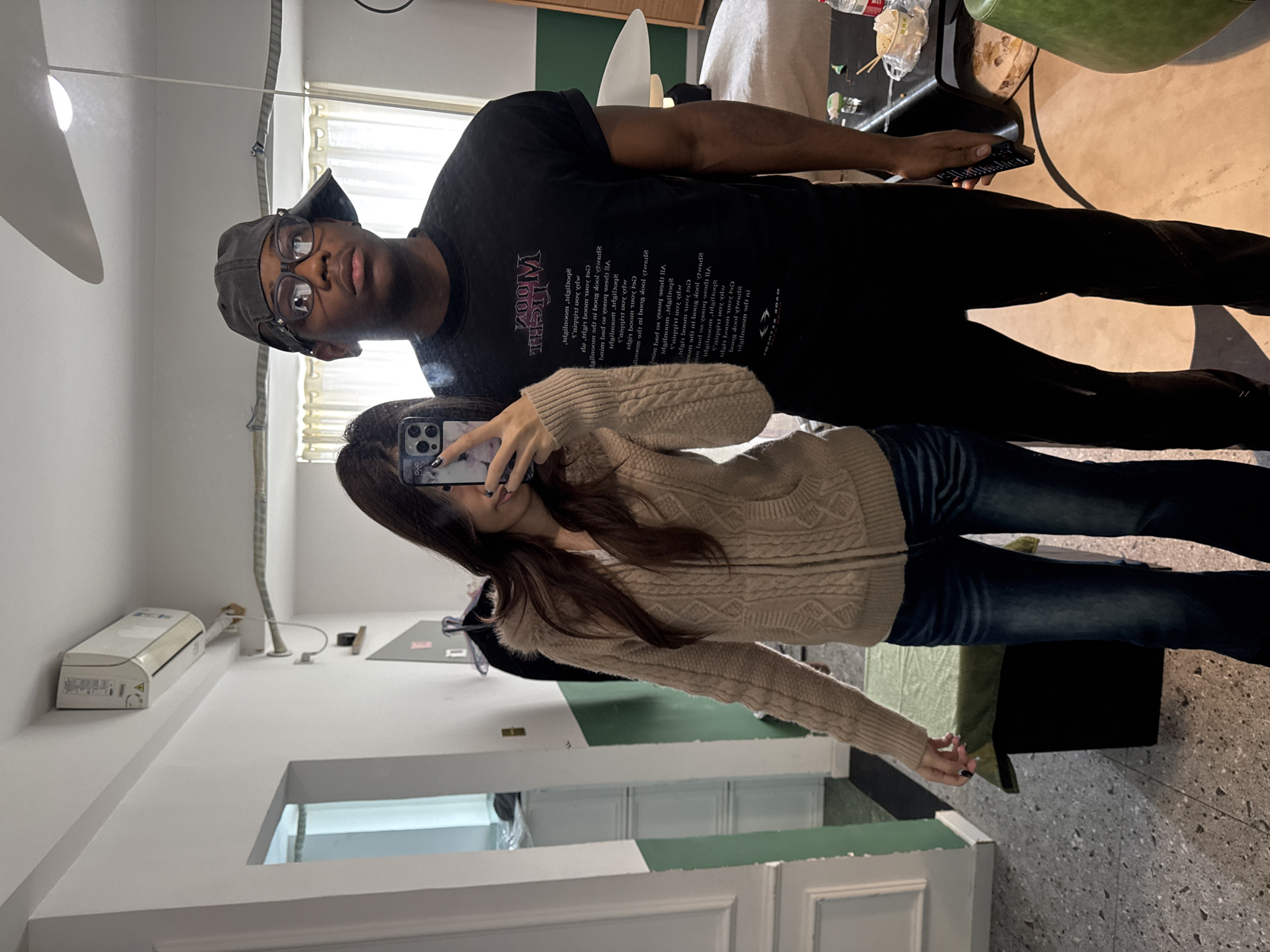 This image depicts two individuals, a female and a male, in an indoor setting within the city of Liangjiang, China. The scene appears to be a mirror reflection.
The female is positioned on the left, holding a dark-colored smartphone, likely an iPhone with a triple camera array, in her right hand, obscuring her face. She has long, dark hair and her fingernails are painted dark. She is wearing a light brown, cable-knit zippered cardigan with a faux fur-trimmed hood over a white garment, and dark blue jeans.
The male is standing to her right, wearing a black baseball cap worn backward, clear-framed eyeglasses, a black short-sleeved t-shirt featuring vertical white text (partially legible as "MOON LIGHT" in mirrored script), and black pants. He holds a black remote control in his right hand. His gaze is directed slightly upwards and to the right.
The environment features white walls with a green wainscoting section below. The floor is light grey terrazzo. A split-unit air conditioner is mounted high on the wall in the upper left, and a white ceiling fan is visible in the reflection above the male's head. To the left, a white-framed doorway opens into another room. Behind the male, a window with white curtains is visible, along with a dark, low table holding a clear water bottle, a small bowl containing food, and chopsticks. A light-colored lamp with a broad, flat shade is partially visible to the far right. A dark green upholstered bench or low table is situated to the left of the female's lower body.