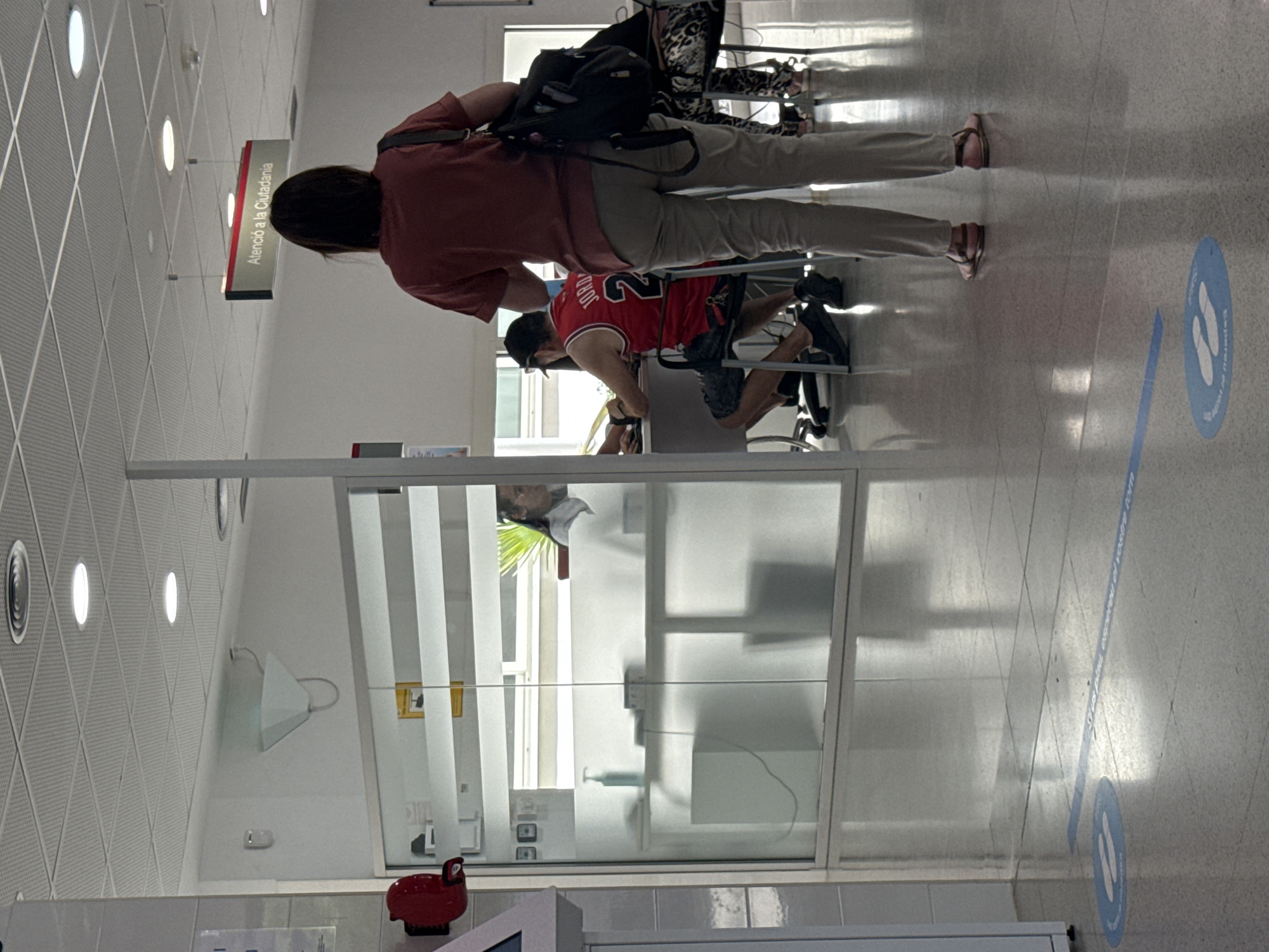 In a public service building, likely a government office or similar establishment in L'hospitalet De Llobregat, Spain, a woman stands facing away from the camera. She has dark hair and is wearing a maroon t-shirt and khaki pants. She carries a dark backpack over her right shoulder. The scene is indoors, with a tiled ceiling featuring recessed lighting. A sign above her reads "Atenció a la Ciutadania," indicating citizen services.
To the left, a glass-enclosed office area is visible. Inside, a person wearing a red basketball jersey with "JORDAN 23" on the back is seated at a desk, facing away from the camera and interacting with someone out of frame. Another person, possibly a woman with glasses and dark hair, is seated behind a counter inside the office. On the counter, a bottle of hand sanitizer and a laptop are present.
The floor is shiny and tiled, with blue social distancing markers in the form of footprints visible on the ground. These markers suggest a queueing system is in place. The overall environment appears clean and organized, typical of a public service waiting area. The lighting is bright and artificial.