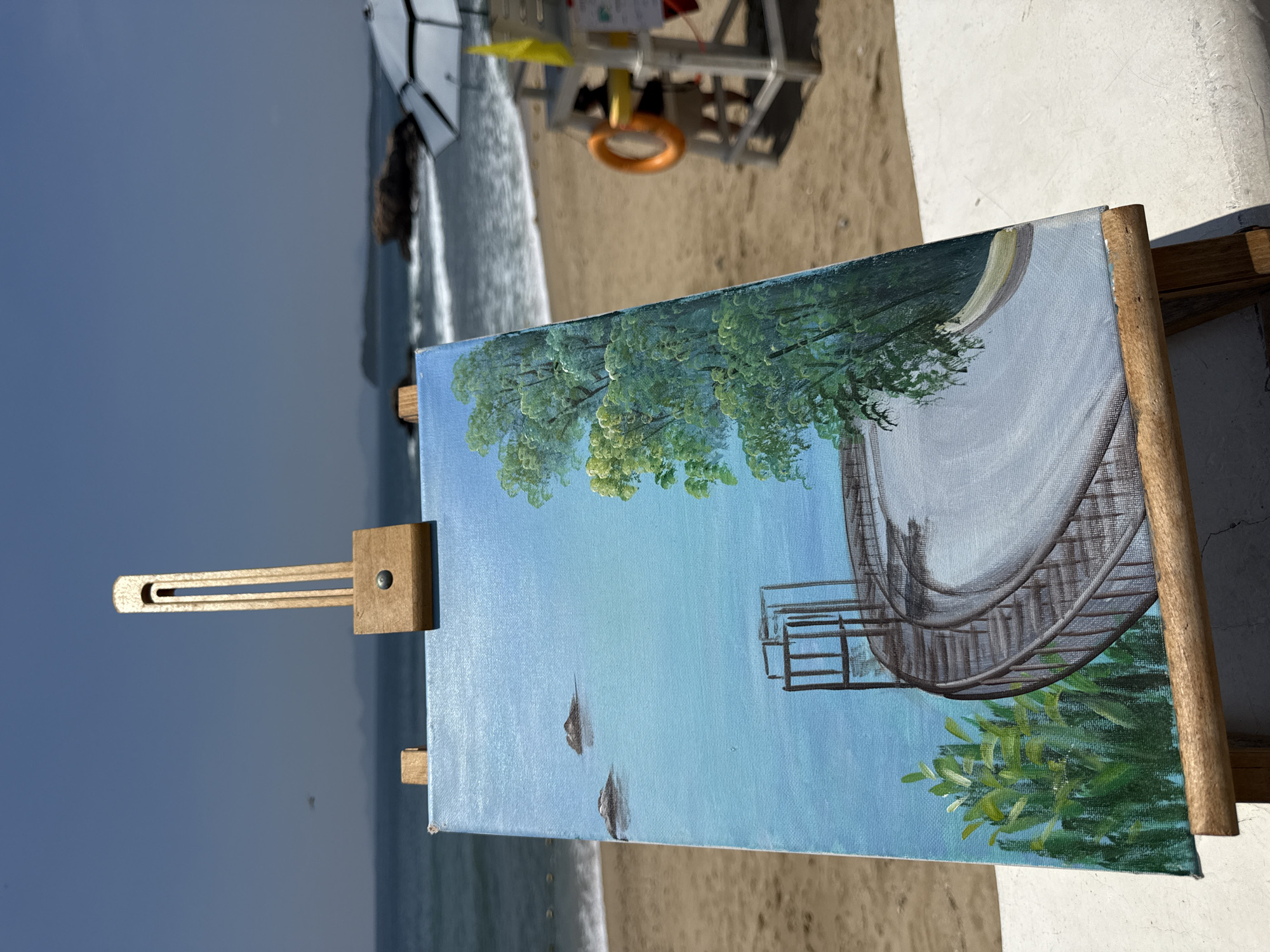 This image depicts an easel with a painting of a seaside scene. The painting shows a calm blue ocean under a clear sky, with two small islands in the distance. A circular pathway with a railing curves into the foreground, bordered by lush green trees. In the background, the actual beach is visible with sand, gentle waves, and a lifeguard stand with a person and a life preserver. The location is identified as Longgang District, China, suggesting this is a view from or near a coastal area within that district. The painting is positioned on the beach, possibly for plein air painting.