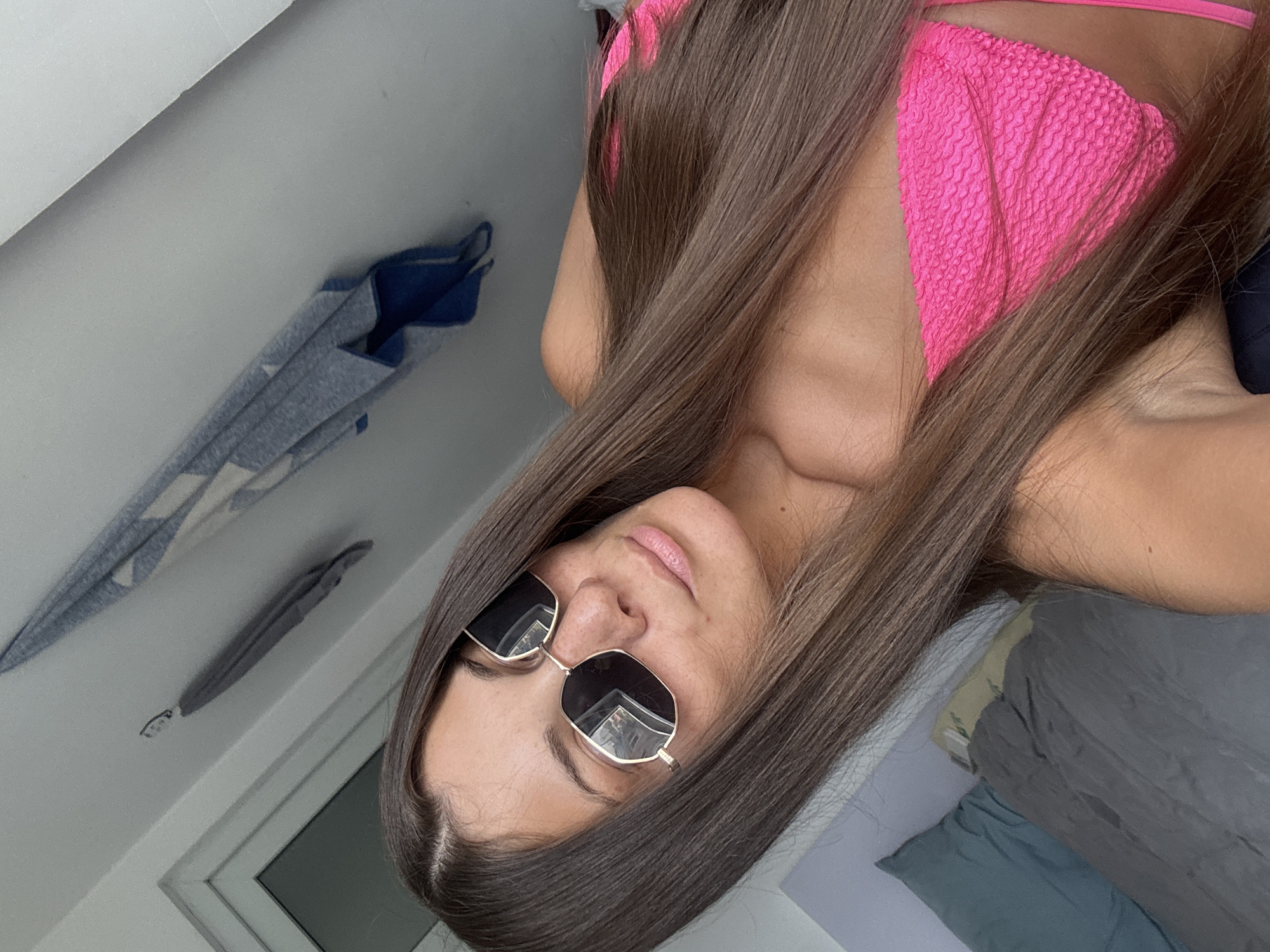 A woman with long brown hair wearing a pink bikini top and sunglasses is the focal point of the image. She is indoors, with a window visible in the background. On the wall, there is a blue and white garment hanging. A bed is visible in the background. There is no information to verify the location of the image.