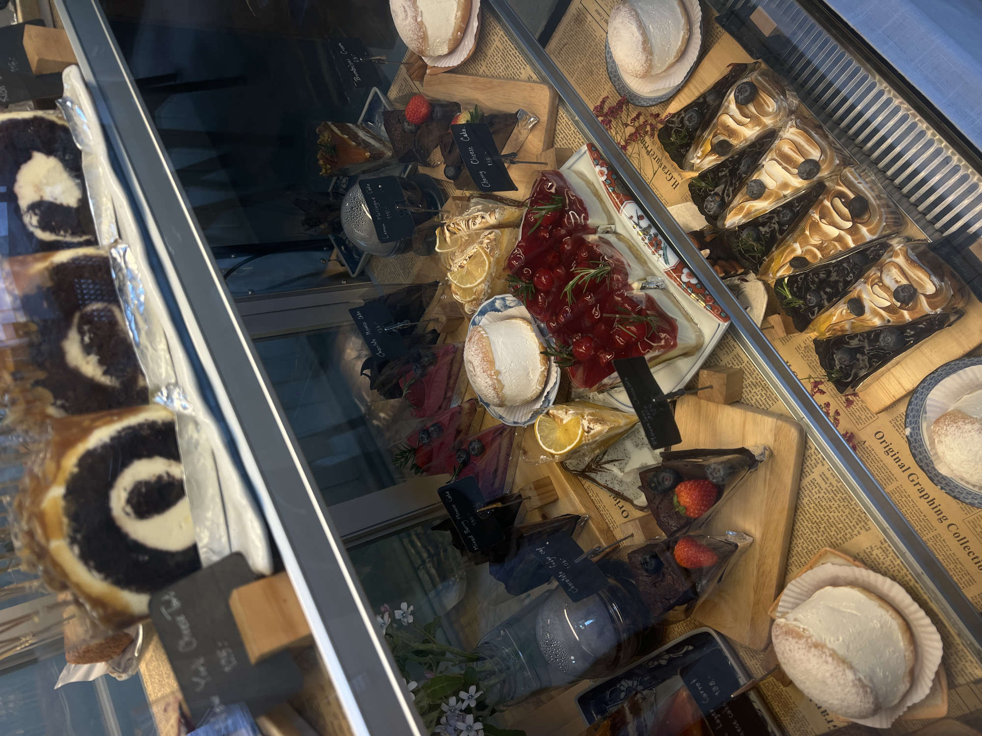 Here is a detailed description of the image:
**Scene:** A display case filled with various desserts.
**Location:** Unknown city, Unknown (indicated by the prices in the signage which may indicate the currency).
**Objects:**
* Assorted cakes: A variety of cake slices including Blueberry Lemon Meringue Cake, Cherry Cheese Cake, Mixed Berry Mousse Cake, Chocolate Mousse Cake, Chocolate Fudge cake, and Layer Carrot Cheese cake.
* Roll cakes: York Cheese Tart.
* Cream-filled buns: Bonboloni Cream.
* Signage: Small black signs with white text detailing the names and prices of the desserts.
* Display case: Glass-fronted display case.
* Decorative Items: Flowers in a vase.
* Newspaper print: Beneath the items are newspaper print.
* Plates and wooden boards
**Description:**
The display case is brightly lit, showcasing a diverse selection of cakes and pastries. The desserts are neatly arranged on wooden boards, plates and newspaper print. The signage provides the name and price of each item.