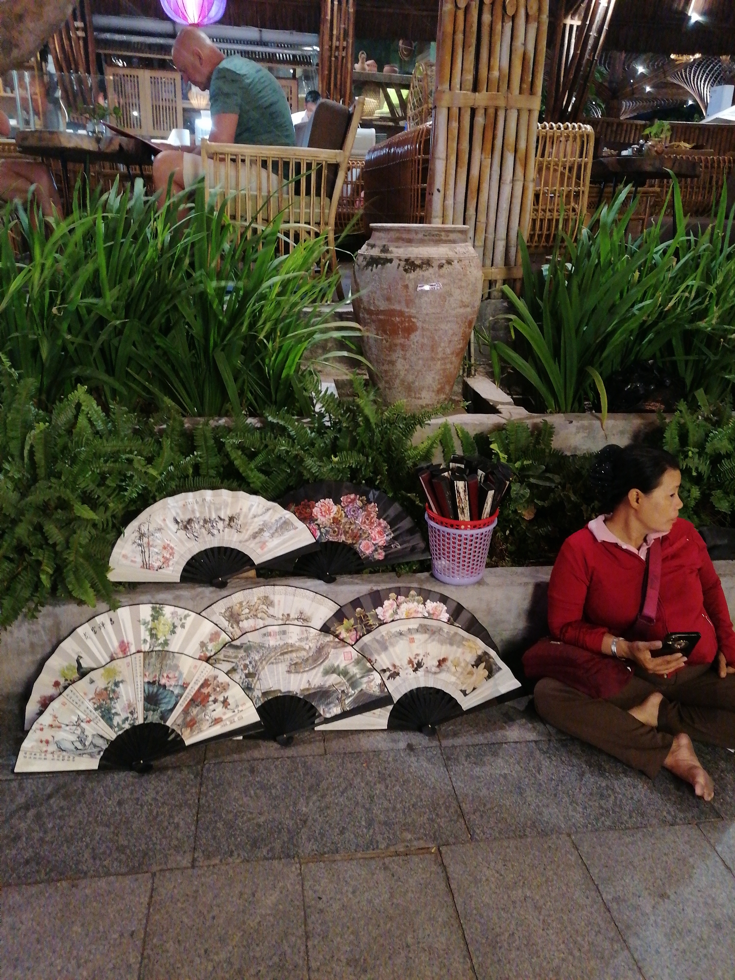 The scene is set in Nha Trang Ward, Vietnam, where a woman sits cross-legged on a tiled surface, surrounded by several ornate hand fans. The woman, dressed in a red jacket and brown pants, is holding a black smartphone in her hands. Beside her is a purple plastic container holding more of the hand fans.
The fans themselves are displayed along the wall behind her, depicting various scenes including landscapes and floral arrangements. Behind the woman and the fans is a raised garden bed overflowing with lush greenery, with a large clay pot situated in the center. In the background, there is outdoor seating at a bamboo-constructed establishment. A man in a green shirt is seen at one of the tables. The setting appears to be a tourist area or market.