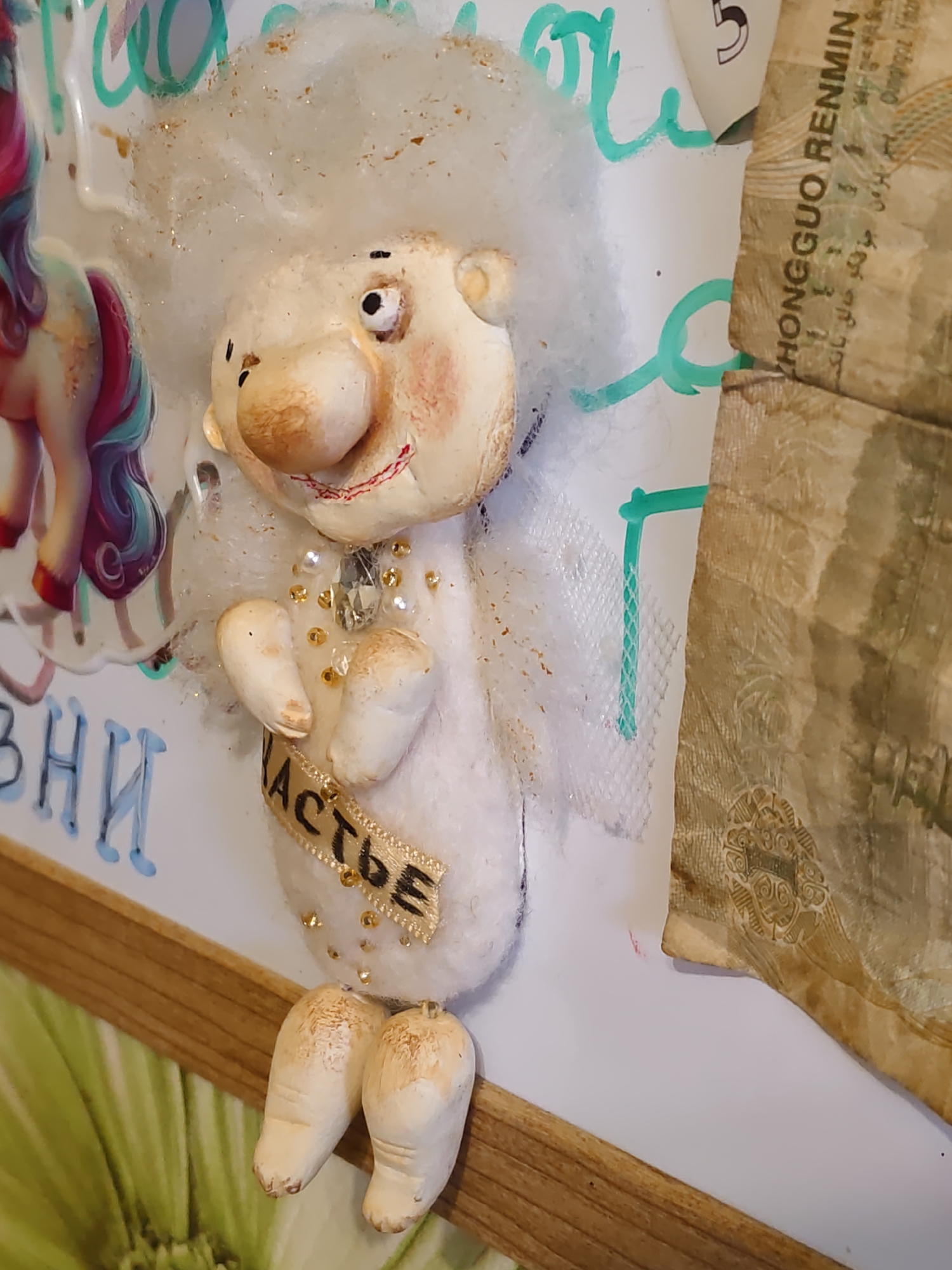 A whimsical, handmade doll with a large, round head and sparse white hair sits in a composition that includes paper money and colorful markings. The doll's face is expressive, with large, prominent cheeks, a bulbous nose, and slightly crooked eyes. It wears a white, fluffy outfit adorned with gold beads and a jeweled pendant. A ribbon with black lettering is wrapped around its torso. To the left of the doll, a colorful depiction of a unicorn is visible. The background features handwritten text in teal and blue, and a portion of aged paper money, possibly a renminbi, is attached to the right side. The scene is framed by a wooden border. The image was taken in Rostov-on-Don, Russia.