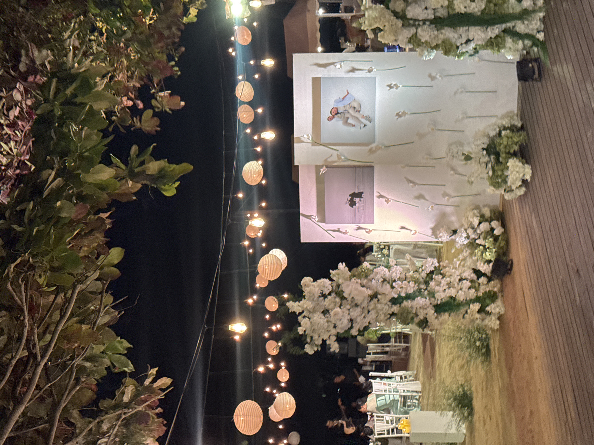 The image captures an outdoor scene, likely at night, in what appears to be a wedding or event setting. The location is potentially in Denpasar, Indonesia, though that's difficult to confirm. The primary focus is a decorated structure or backdrop, possibly a wedding arch, adorned with white flowers and greenery. On the backdrop are two framed photographs. String lights, some with paper lanterns, hang in the foreground, creating a festive atmosphere. To the left, there's a tree with leaves visible. In the distance, chairs are arranged, likely for guests. There are also people visible. The ground appears to be made of wood planks.