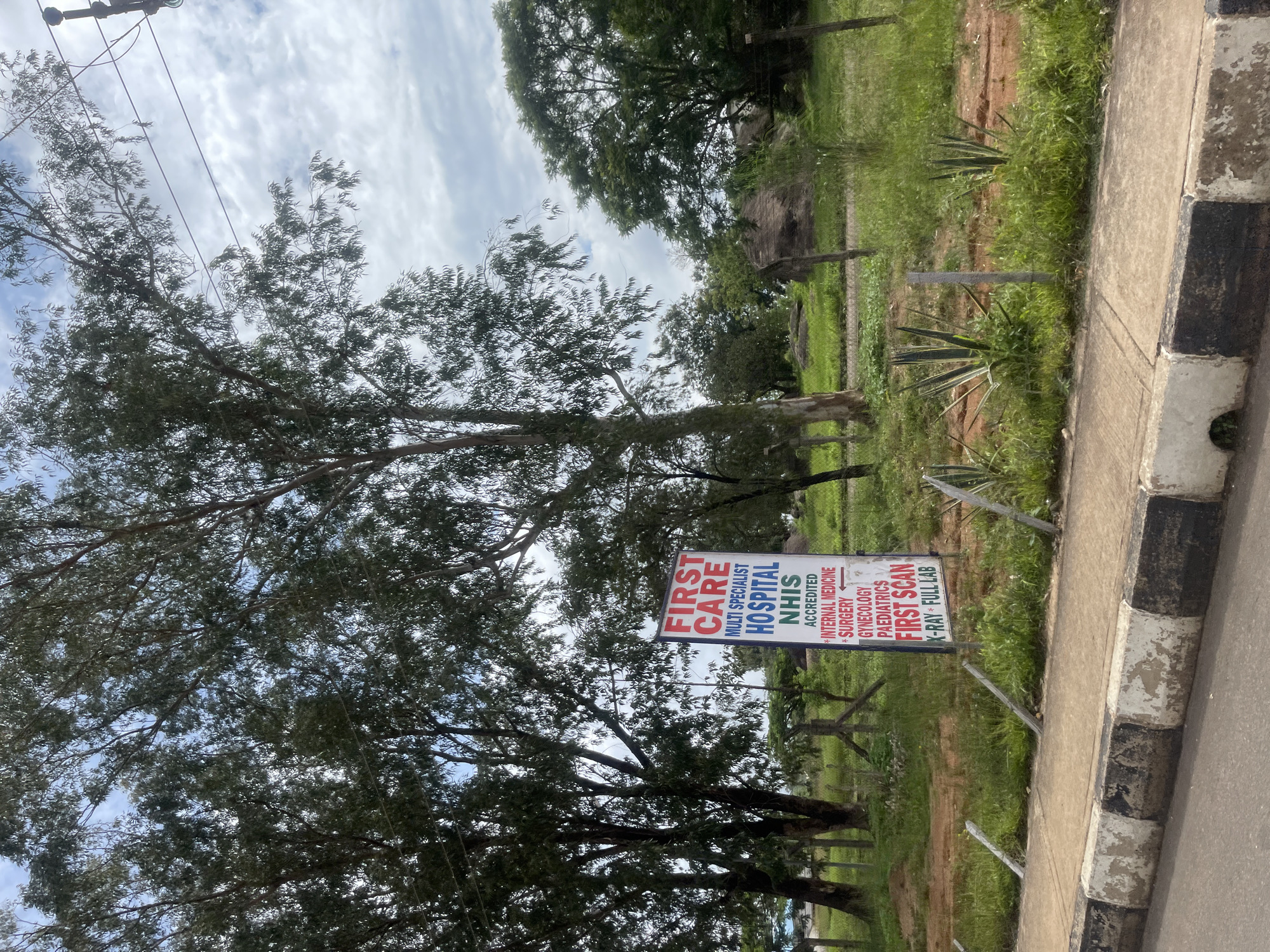 A vertical, full-frame, outdoor, eye-level shot shows a sign for "First Care Multi Specialist Hospital NHIS" situated in a rural landscape with scattered trees and green grass. The sign, positioned in the middle ground, is a white rectangular board with red and blue text. The red text at the top reads "FIRST CARE" in large capital letters. Below this, in blue capital letters, is "MULTI SPECIALIST HOSPITAL NHIS". Further down, smaller white text lists services: "ACCREDITED", "INTERNAL MEDICINE", "SURGERY", "GYNECOLOGY", "PAEDIATRICS", "FIRST SCAN", and "X-RAY FULL LAB".
In the background, tall, slender trees with dark green foliage dominate the left and right sides of the frame. The sky above is a mix of blue and white clouds. The ground around the sign is covered in green grass with patches of reddish-brown earth. A rough, dark-colored structure, possibly a thatched roof dwelling, is visible in the far right background. A wire fence runs horizontally across the mid-ground, separating the grassy area from the foreground.
The foreground features a paved sidewalk with a black and white painted curb, showing signs of wear and tear. The overall scene suggests a location in or near a semi-urban or rural area of Bar Jirgi Summa, Nigeria, given the presence of the hospital sign and the natural environment. There are no people or animals visible in the image.