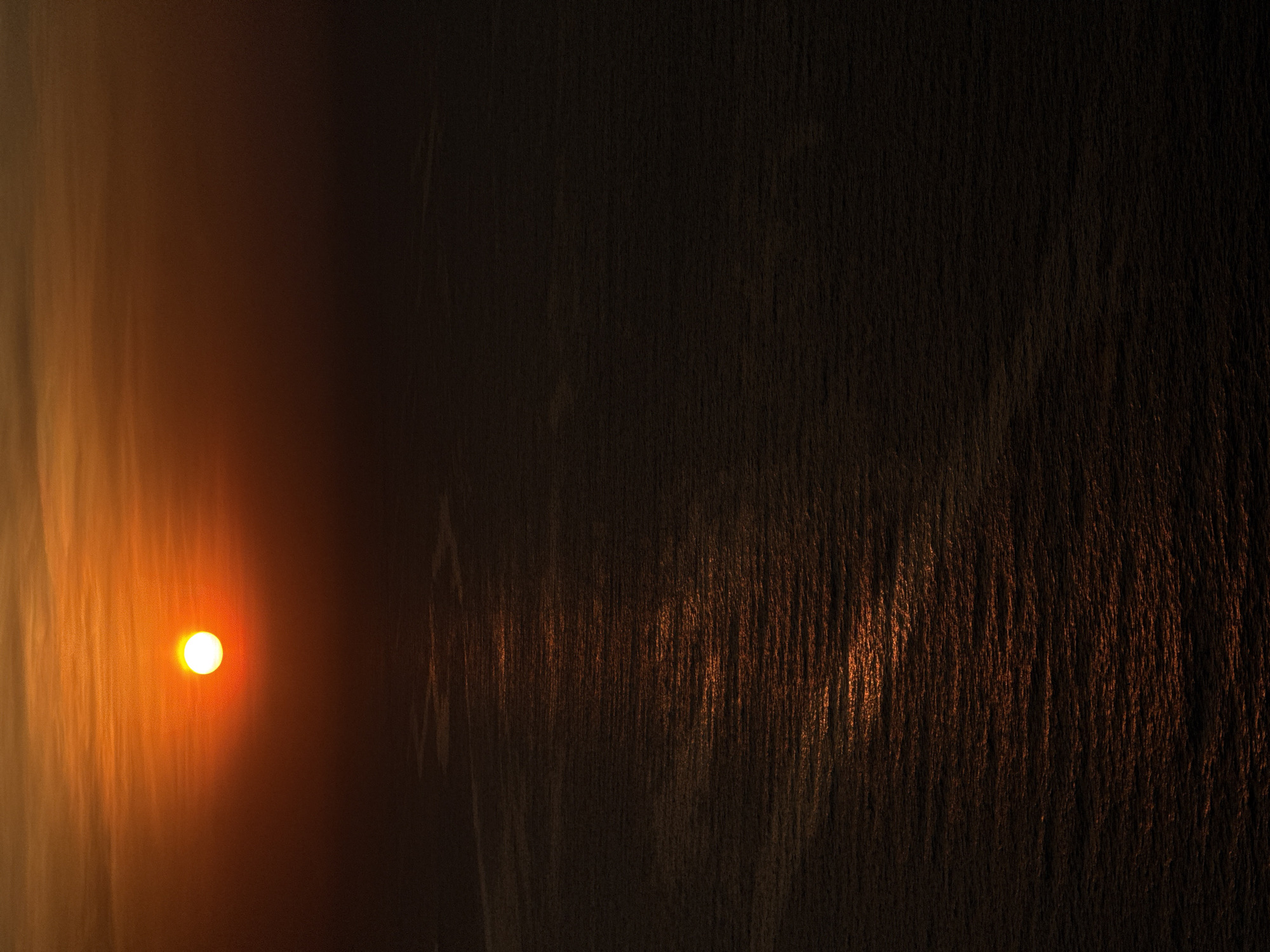 The image depicts a breathtaking sunrise or sunset over a vast body of water, likely off the coast of Đà Nẵng, Vietnam. The bright, white-yellow sun is a prominent feature, positioned towards the upper-left if the image is viewed in a landscape orientation (rotated 90 degrees counter-clockwise from the original). Its intense glow bathes the surrounding sky in warm hues of orange, red, and brown, gradually fading into darker tones further from the light source.
The surface of the water, which covers the majority of the frame, intricately reflects the sun's light. These reflections appear as shimmering, elongated streaks of orange and deep brown, suggesting a textured or rippled water surface. The light creates an effect resembling vertical brushstrokes across the water, particularly noticeable on the right side of the image where the reflections are more defined against a darker backdrop. The areas of water not directly illuminated by the sun's path are deep brown to almost black, emphasizing the stark contrast with the brilliant sun.
No people or man-made objects are visible, allowing the natural spectacle of the celestial event to be the sole focus. The scene evokes a sense of tranquility and the majestic beauty of nature at dawn or dusk. The clear visibility of the sun indicates fair weather conditions. There is no visible text in the image.