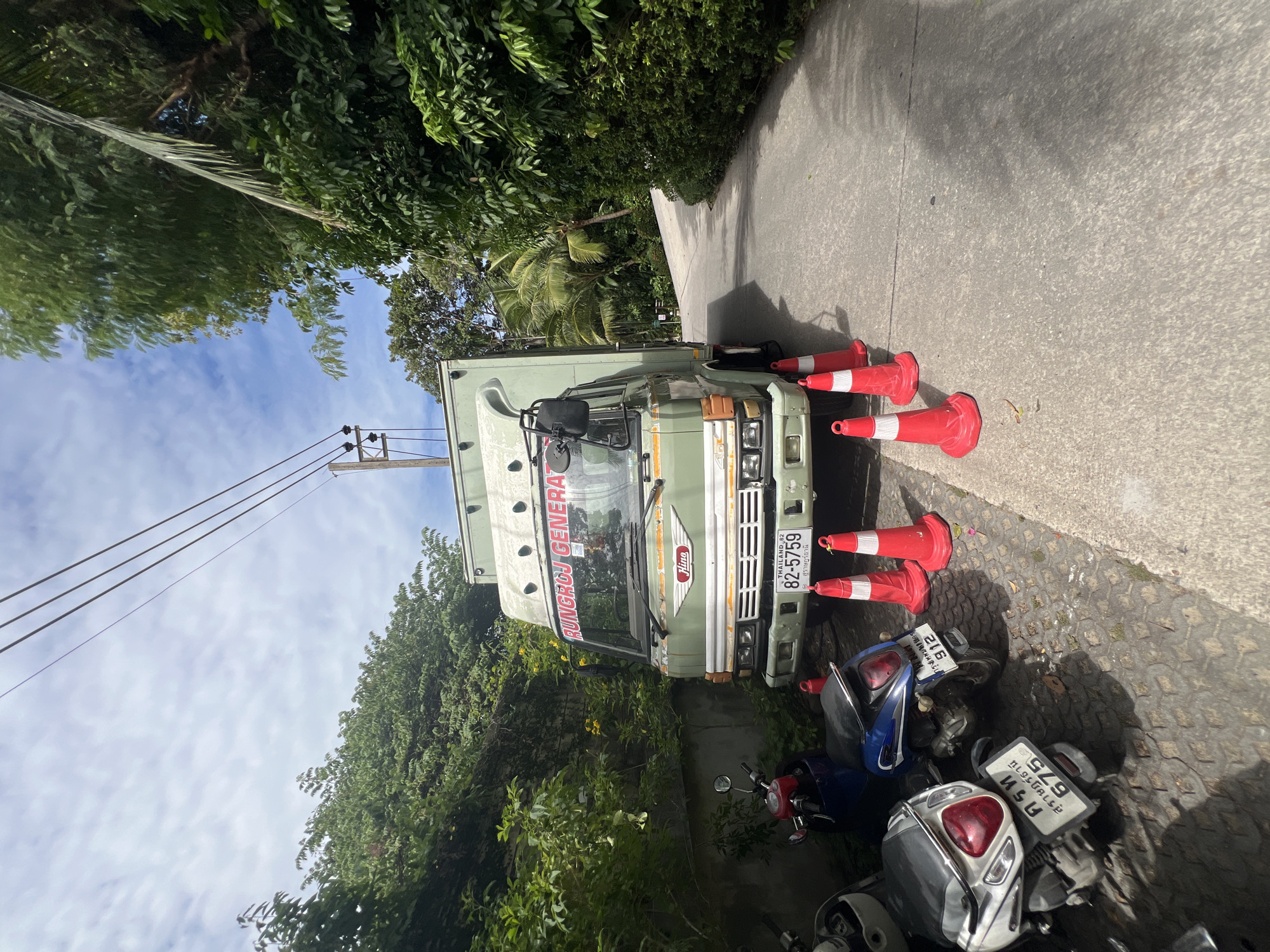 A light green Hino truck is parked on a paved road next to a lush green embankment. The truck has "RUNGROY GENERAL" printed in red on its windshield. Several red traffic cones are placed in front of the truck. In the foreground, two scooters are parked on a cobblestone surface to the left of the truck. Power lines are visible overhead against a blue sky with scattered clouds. The road appears to be a private drive or a road within a tropical area, given the dense vegetation.