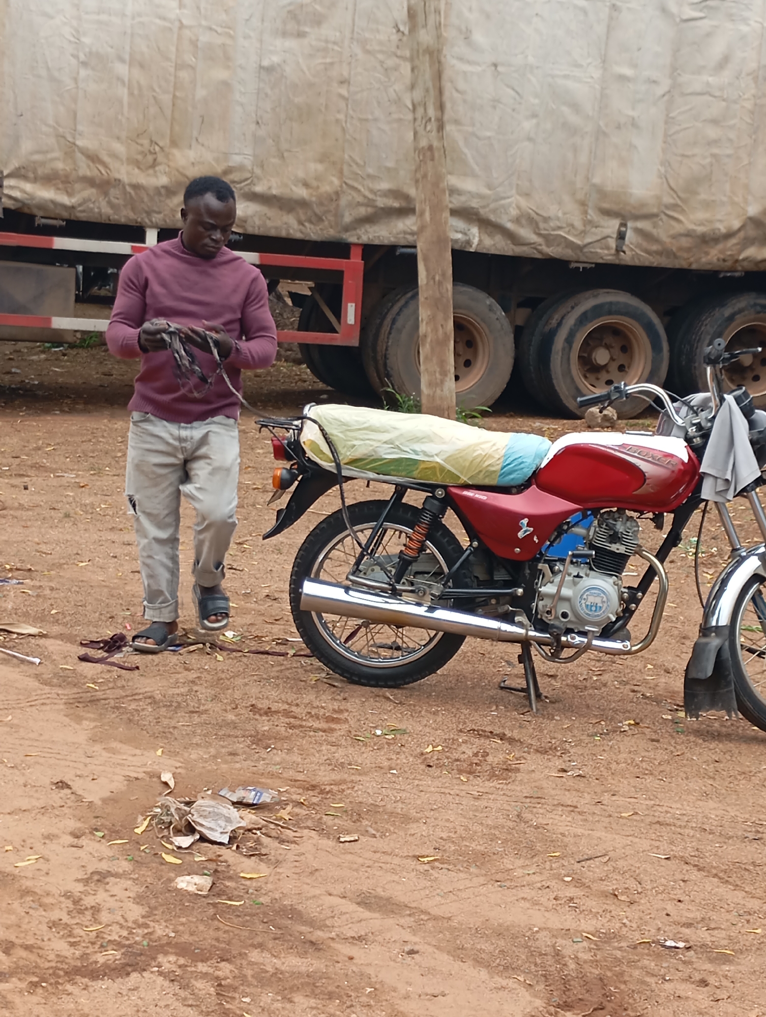 A Black man in a maroon long-sleeved shirt and light gray, ripped jeans stands in front of a large truck in Shendam, Nigeria. He is holding a bundle of dark-colored, thin ropes in his hands and looking down at them. A red and white motorcycle with a light green tarp covering its rear is parked to his right. The ground is composed of reddish-brown dirt with some scattered leaves and debris. In the background, the side of the truck is visible with multiple tires. The scene appears to be outdoors, possibly a roadside or a yard.