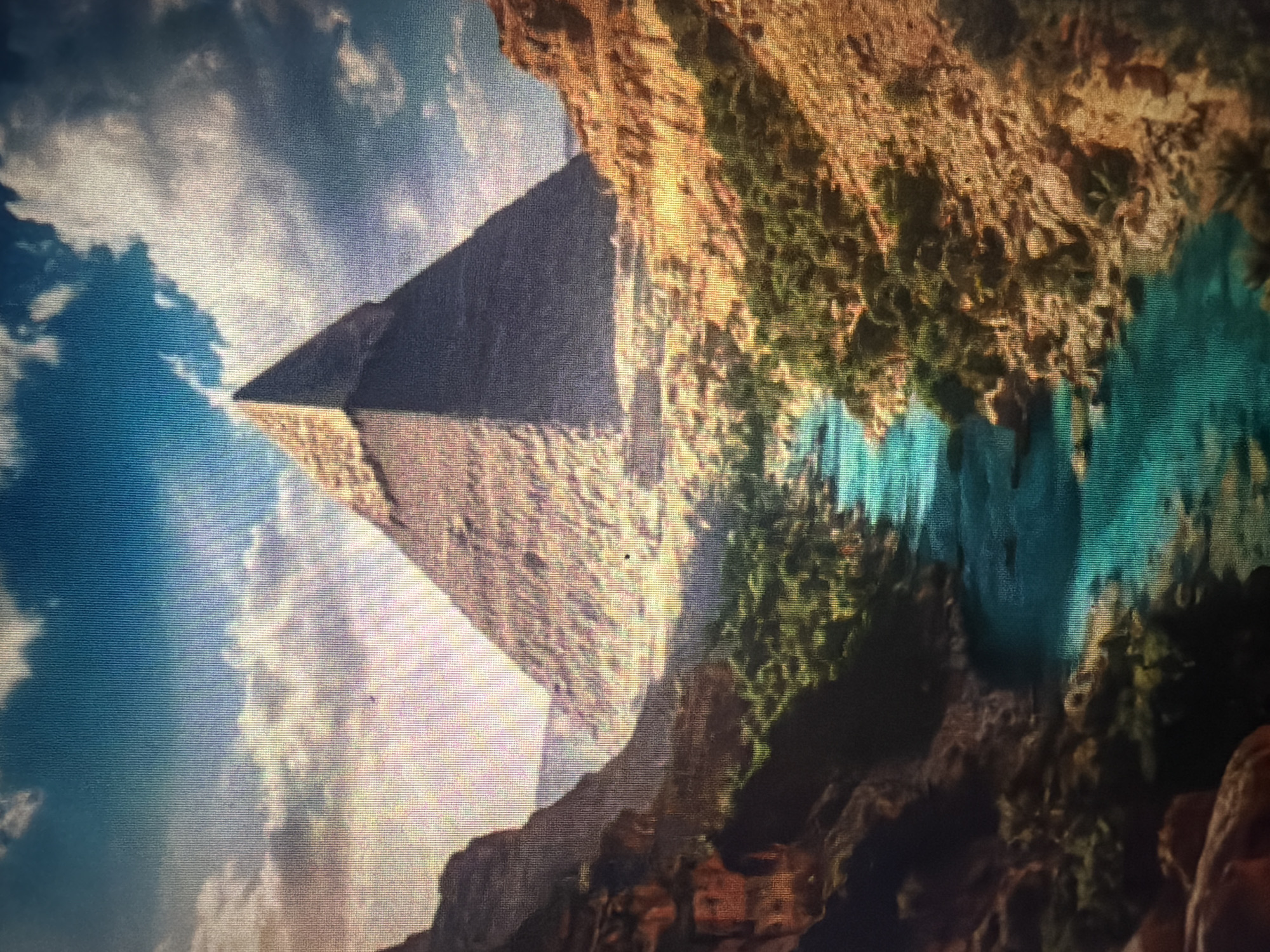 The image depicts a surreal landscape featuring a massive, ancient-looking pyramid situated within a canyon. A vibrant turquoise river flows through the canyon floor, flanked by lush green vegetation on its banks and the rugged, ochre-colored rock formations of the canyon walls. The sky above is a dramatic expanse of blue with scattered white clouds, illuminated by sunlight that casts a warm glow on the scene. There are no people visible in the image. The location in Baan Mae Nam, Thailand, is not depicted or suggested by any elements in the photograph.